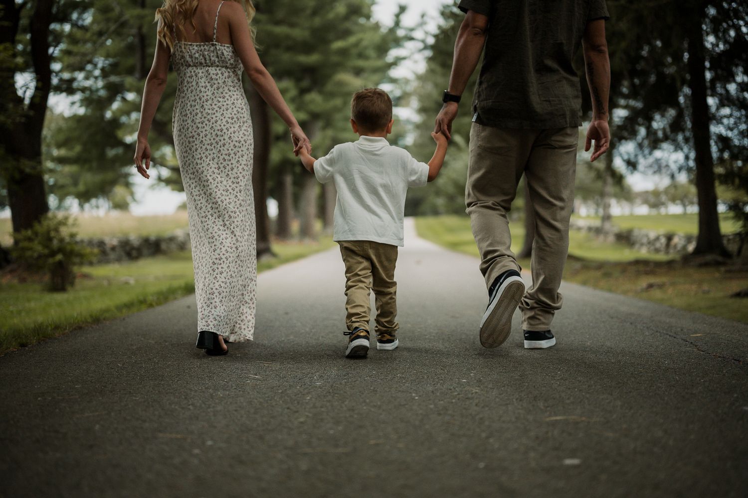Family of three walking hand-in-hand down a tree-lined path, viewed from behind on a cloudy day.