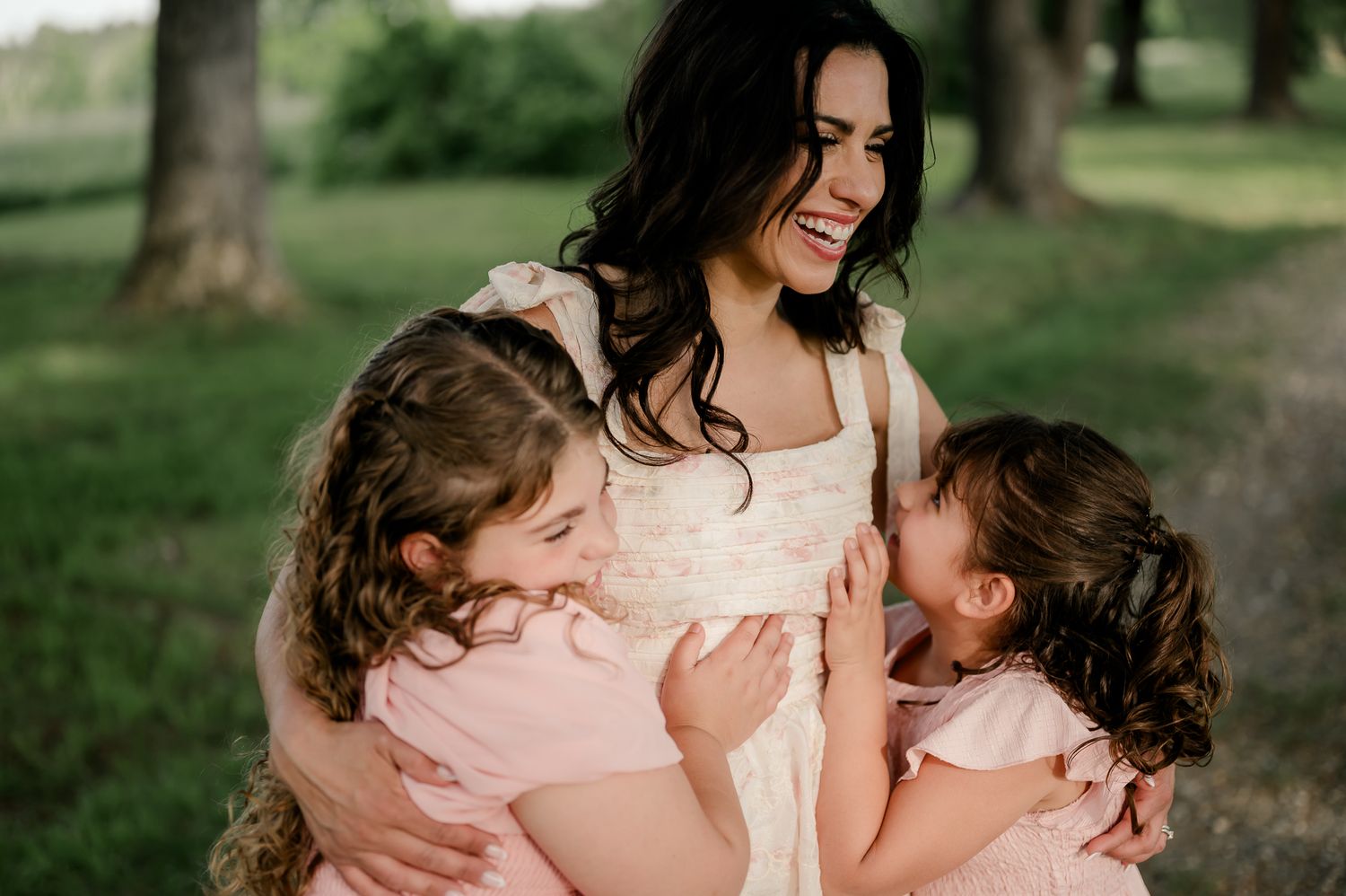 A laughing mother in a cream dress is hugged by two young girls in pink dresses.