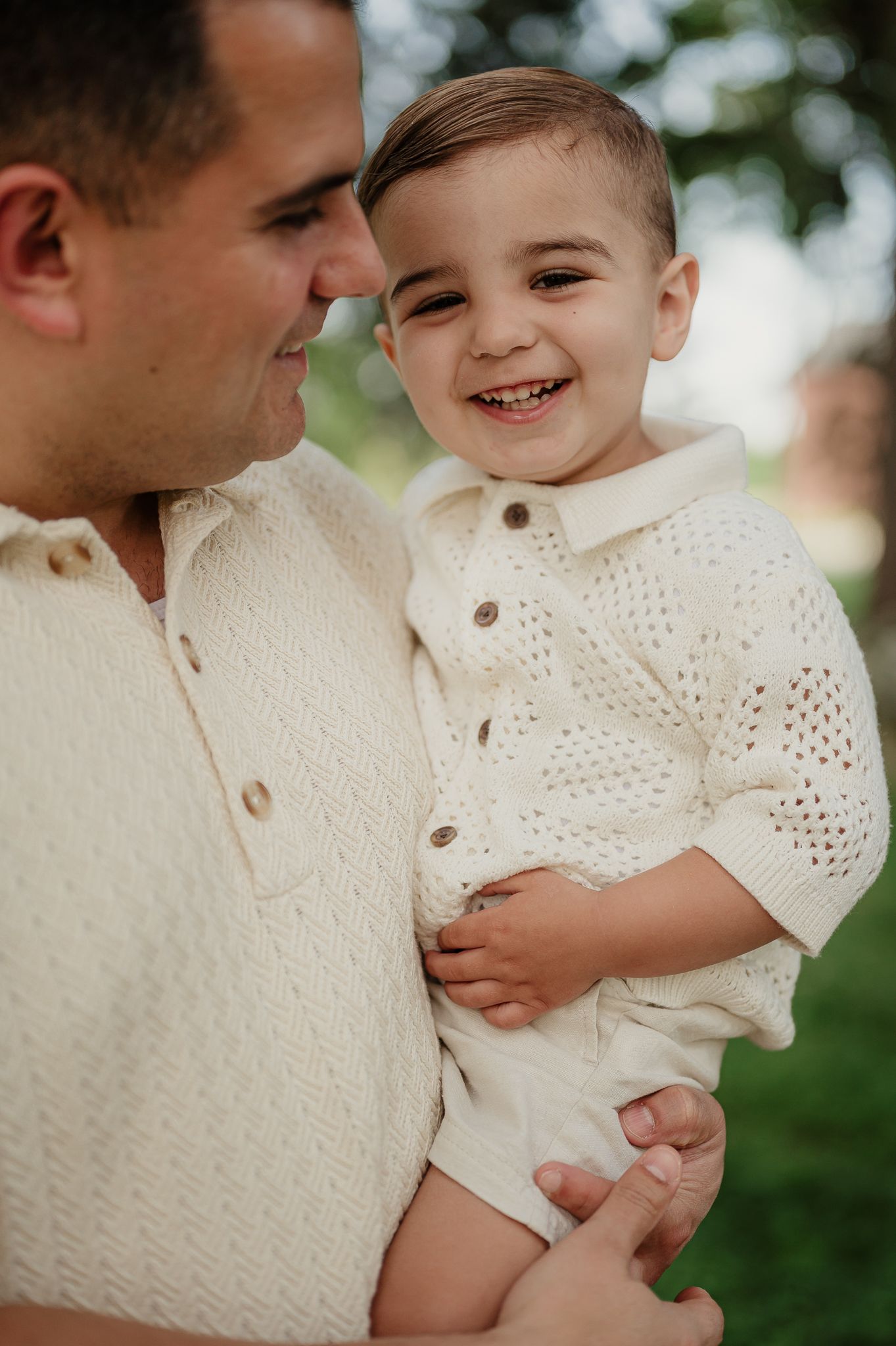A smiling toddler in a cream knit outfit being held by a man in a matching sweater outdoors.