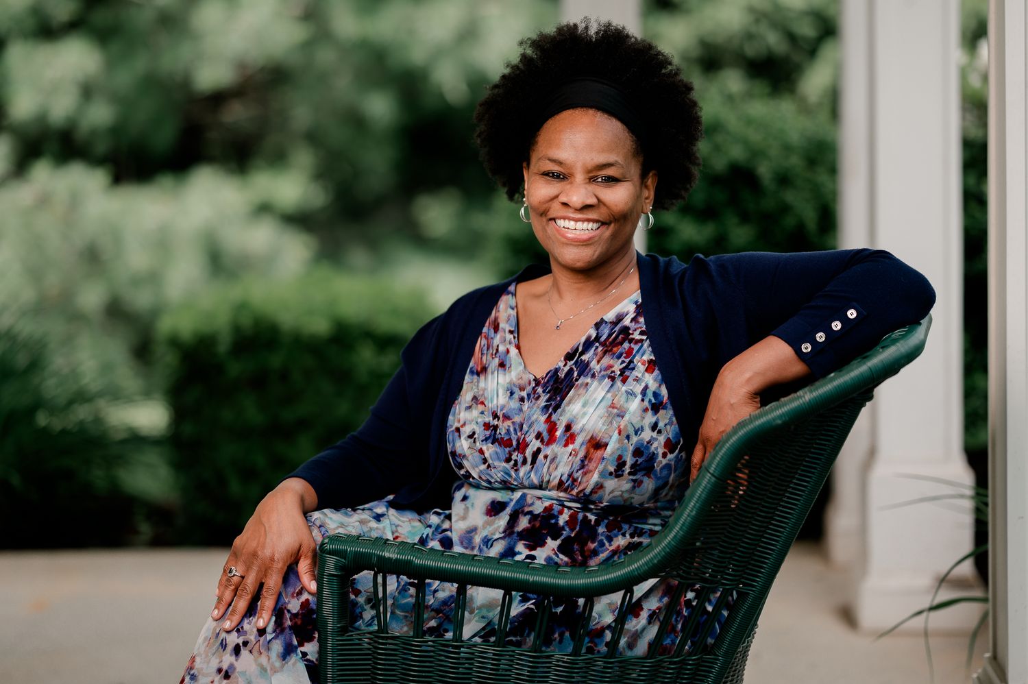 Smiling woman in floral dress relaxing in green wicker chair outdoors among trees.