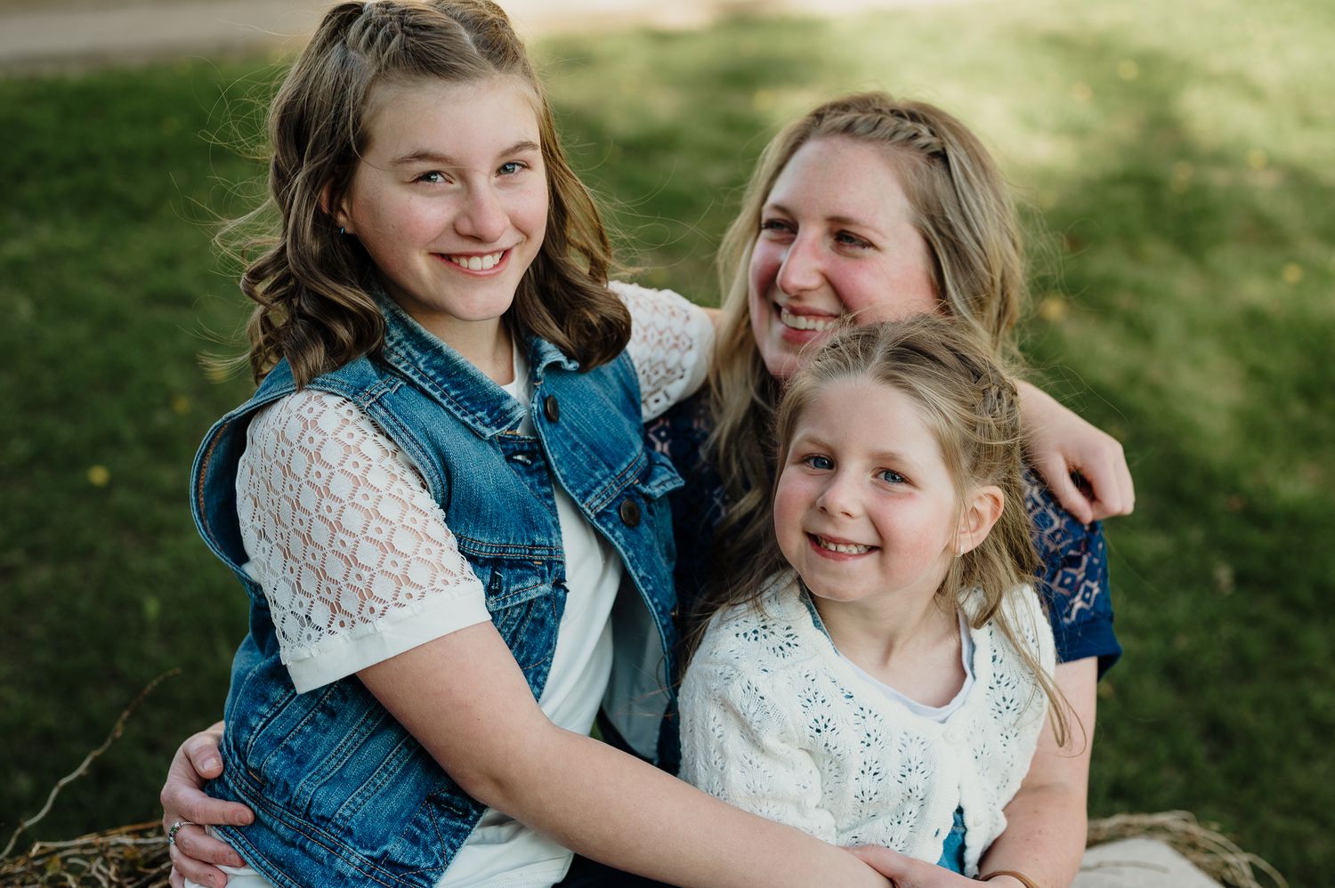 Three smiling girls pose together outdoors on grass, wearing white tops and a denim vest in a warm family portrait.