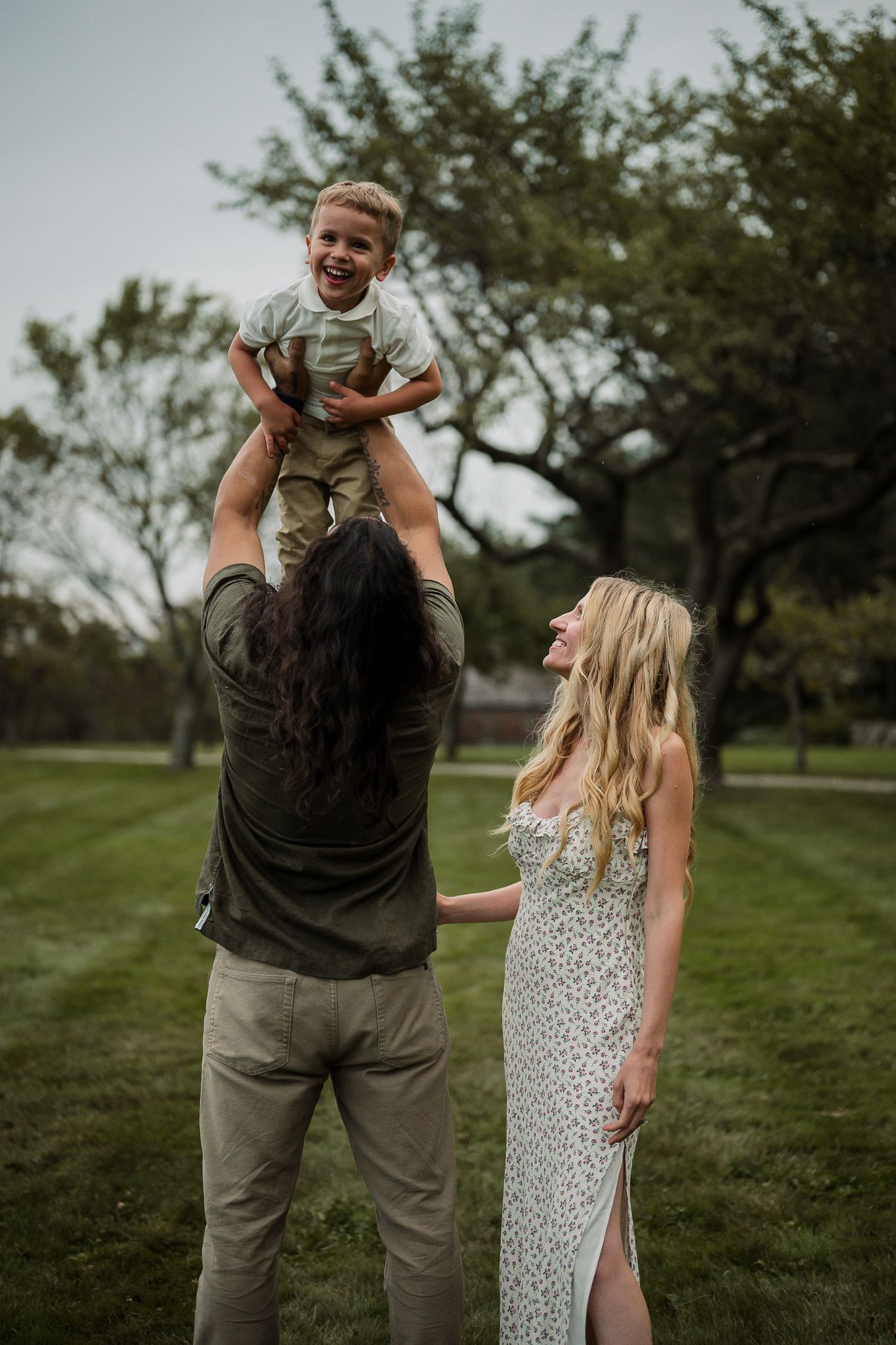 A joyful family moment as a father lifts his laughing toddler son high in the air while mother watches in a park.