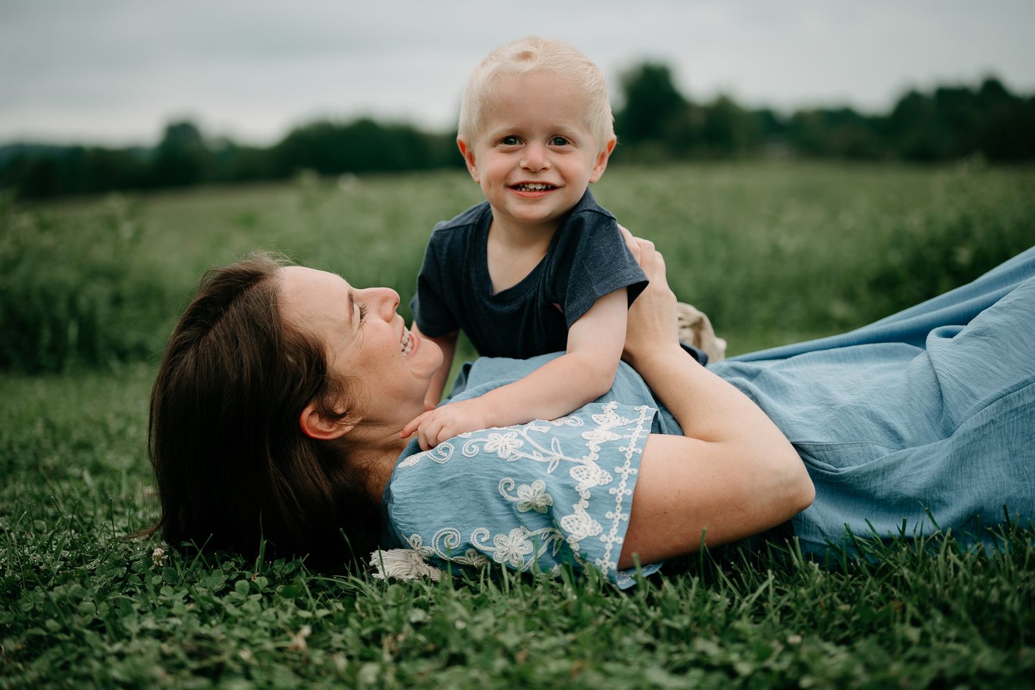 A mother lies on green grass laughing as her smiling toddler boy plays on top of her outdoors.