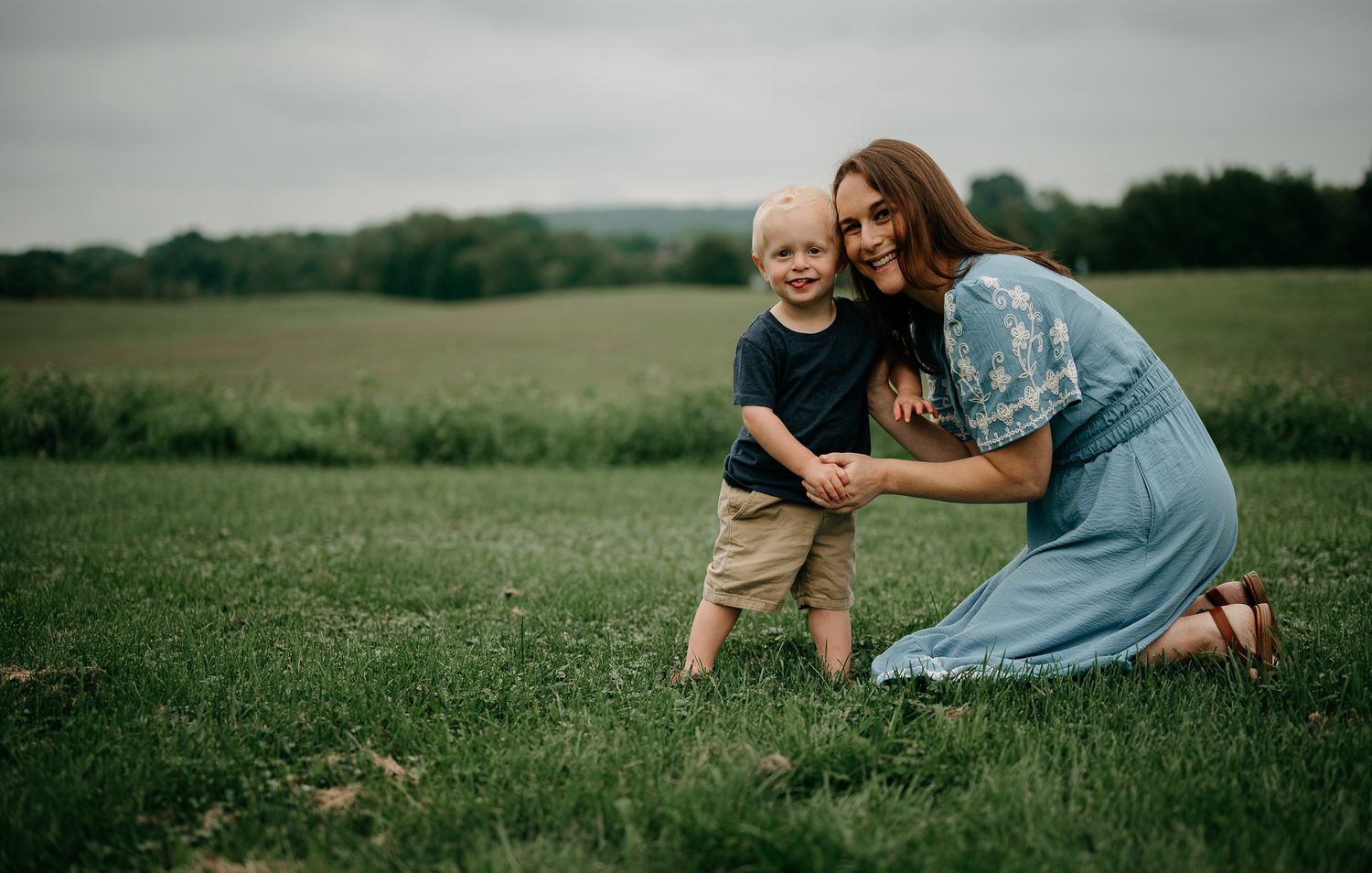 A mother and young toddler boy pose together smiling in a lush green field during an outdoor family photo session.