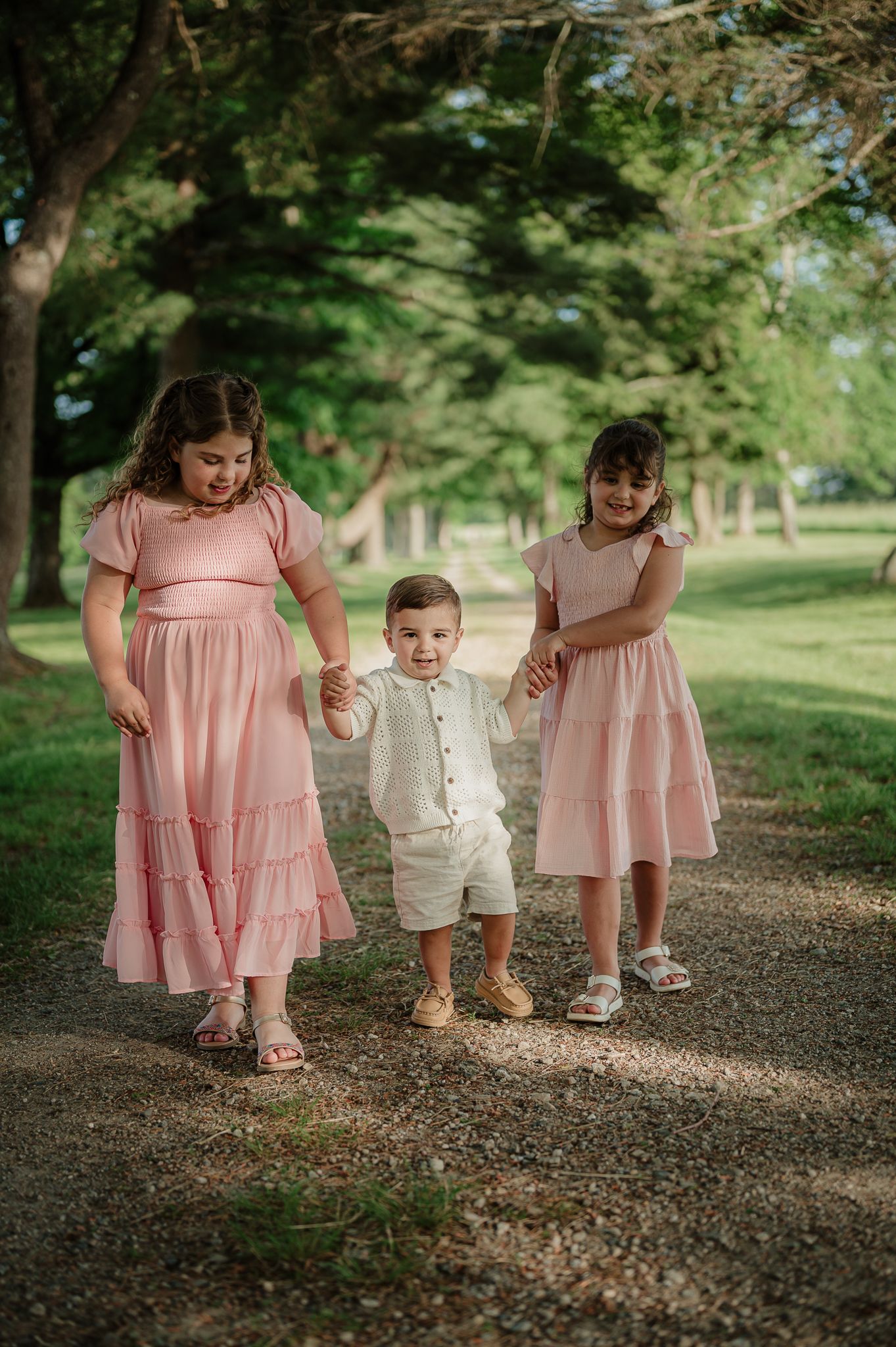 Three children in pink dresses and white outfit walk hand-in-hand on a gravel path surrounded by lush green trees.
