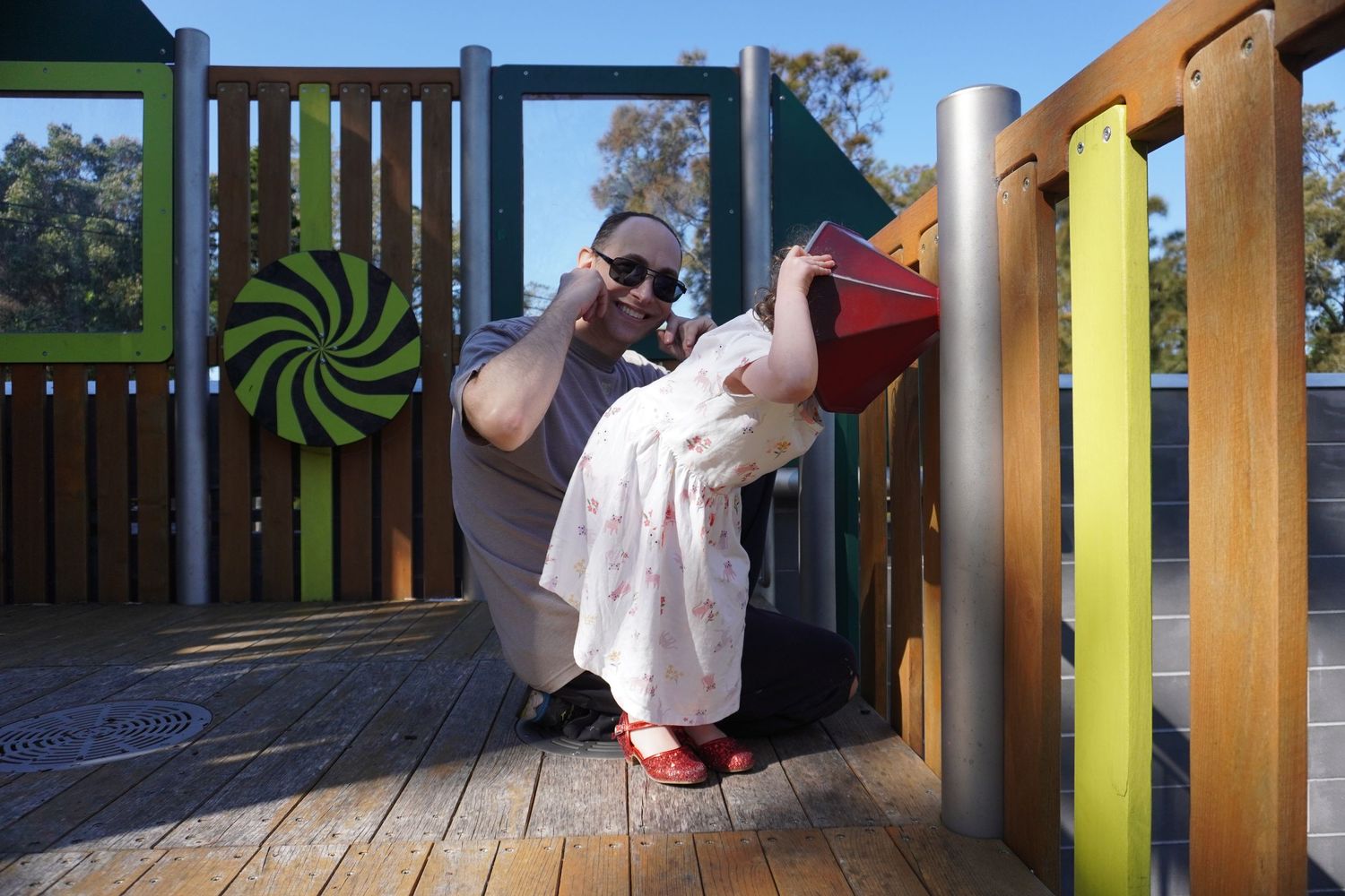 A little girl with red sparkly shoes is at a playground with her father. He blocks his ears & smiles at the camera as she yells into a pipe.