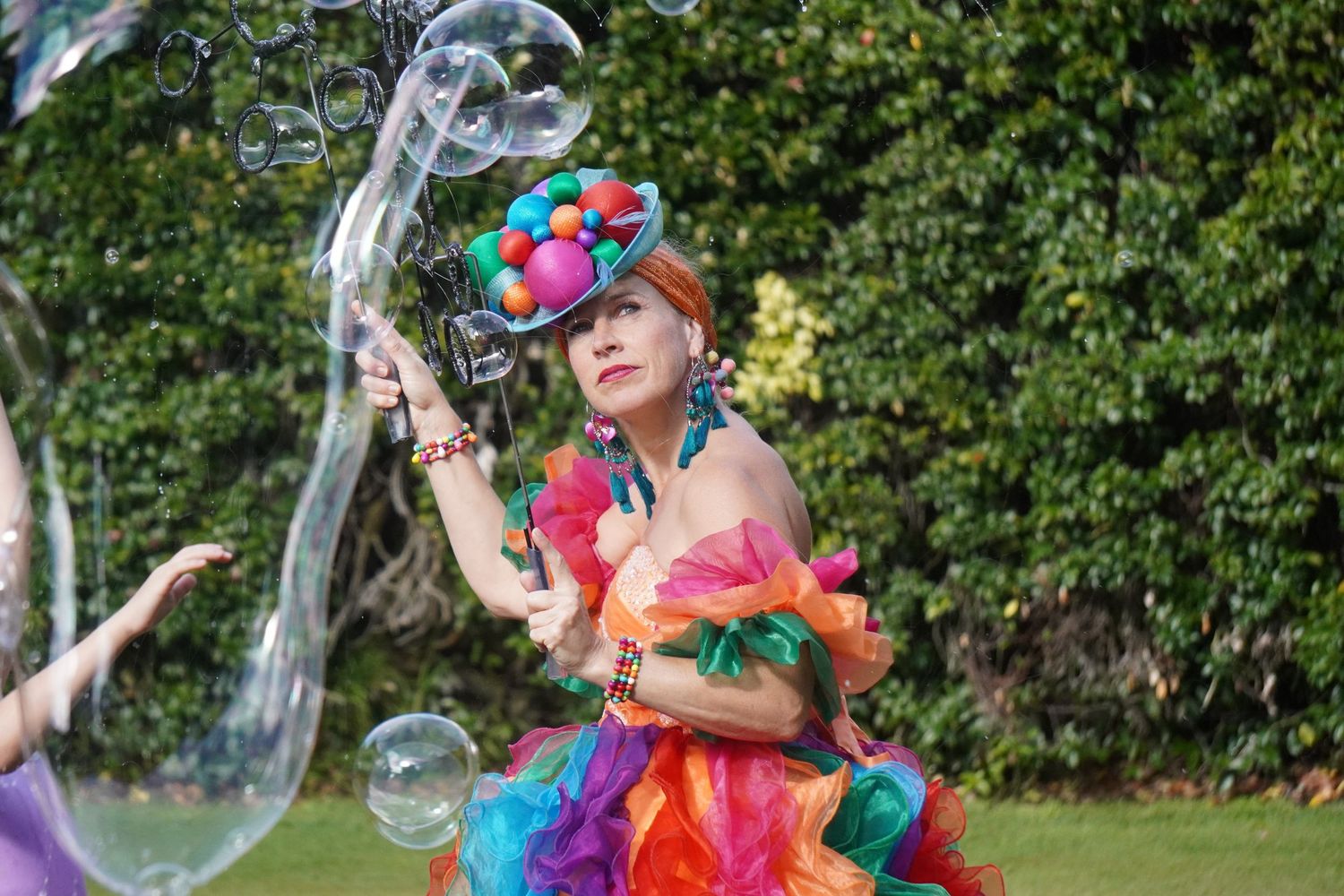 A party entertainer wearing a colourful dress covered in balls makes bubbles. She is looking into the air watching her bubbles float away.