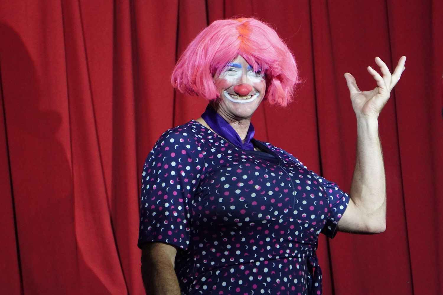 A clown posed and smiling in front of a red curtain. He is wearing a short pink wig, dark blue dress with colourful polka dots, and make up.