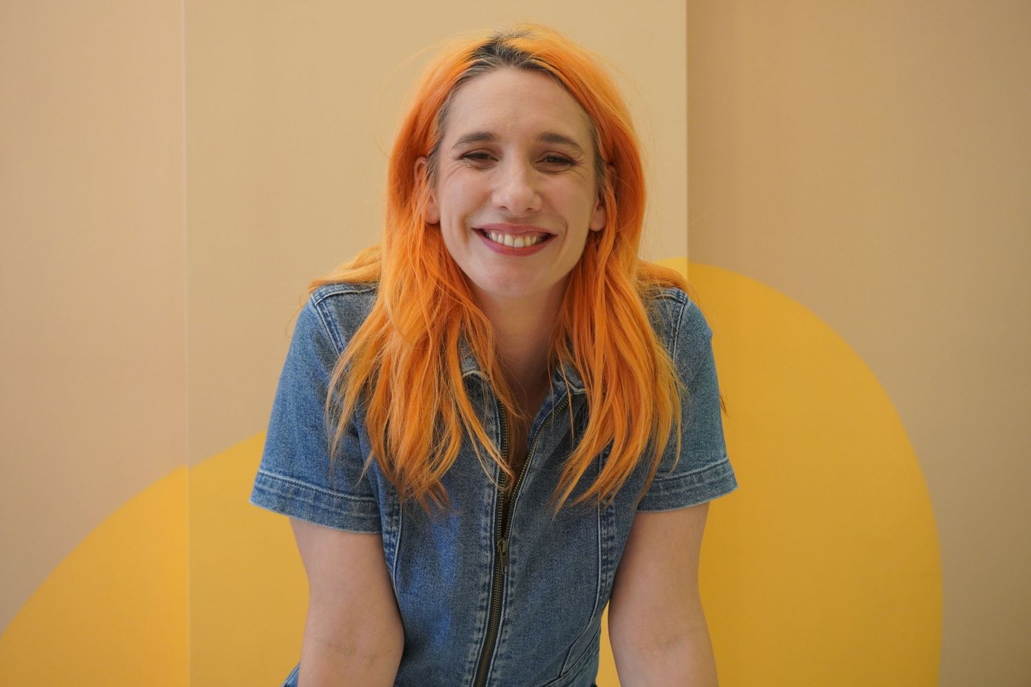 A woman with long straight hair dyed orange smiles at the camera. She is wearing a blue denim jumpsuit.