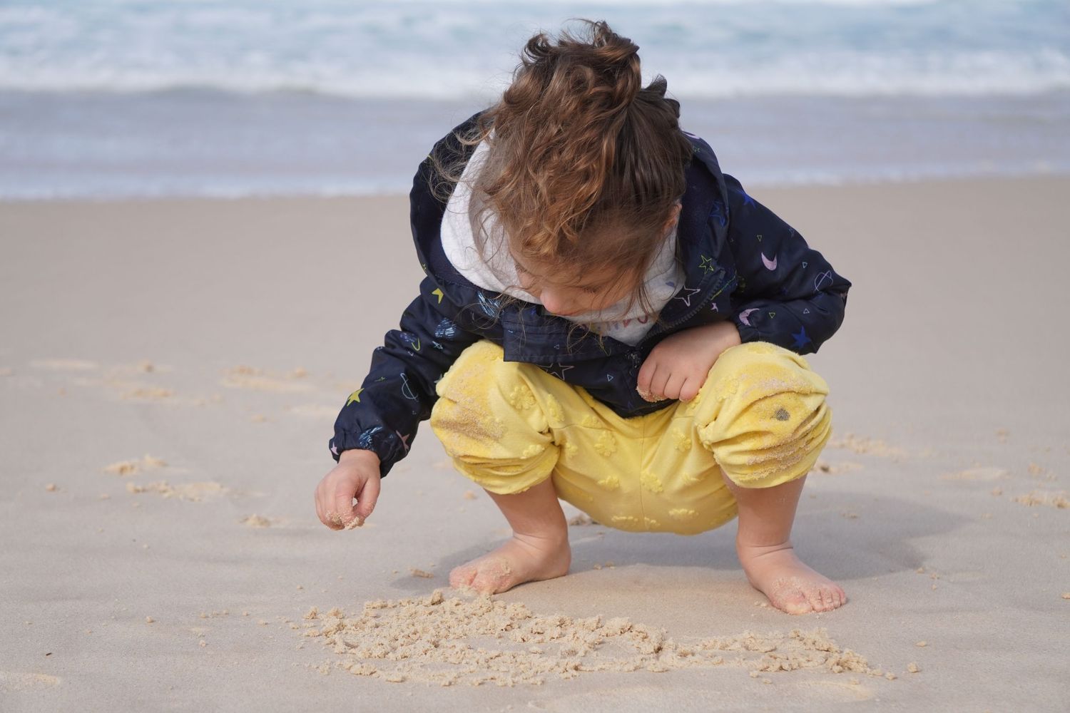 A little girl with brown curly hair at the beach. She is squatting in the sand, looking down, and using a finger to draw in the sand.