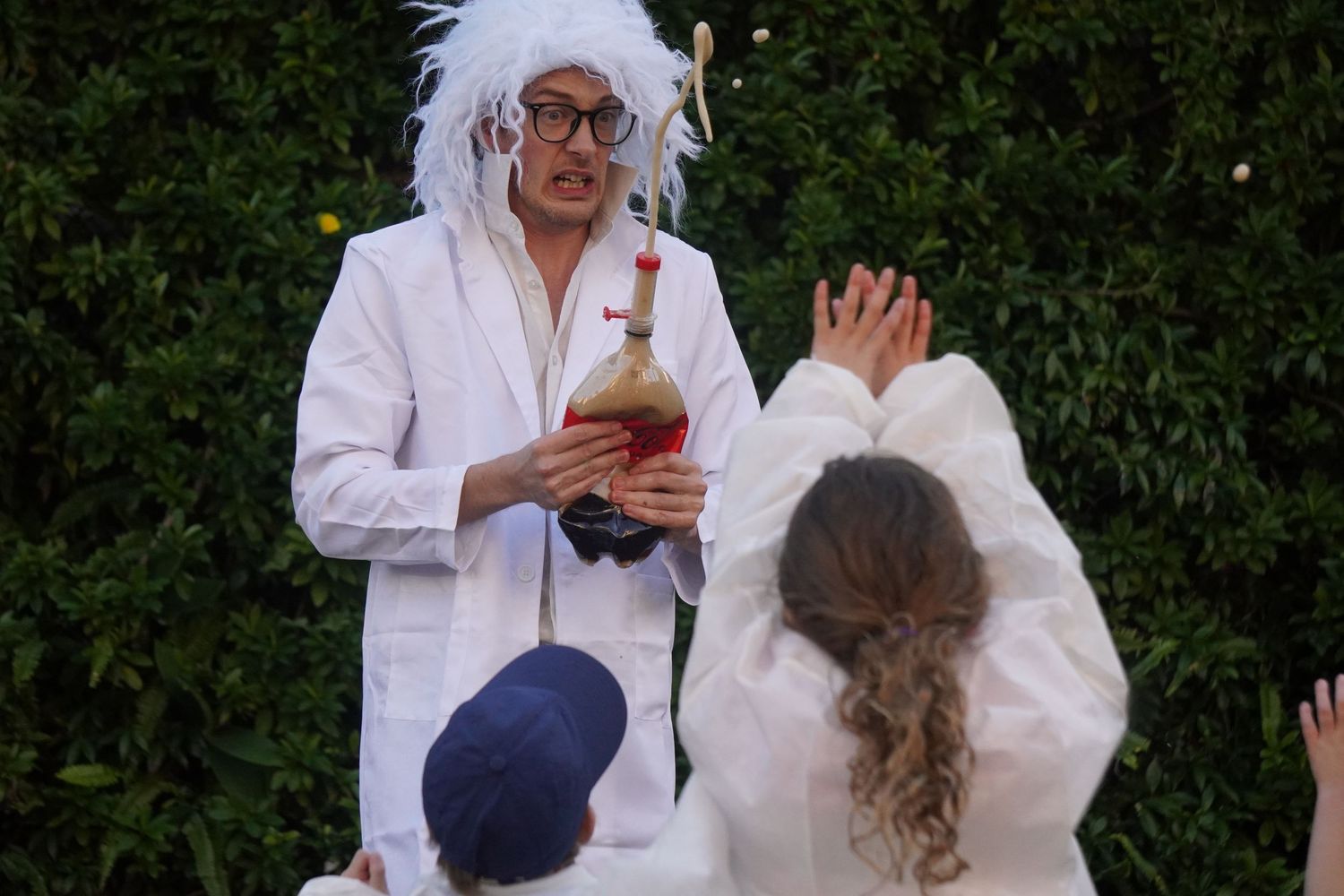 A kids party entertainer is wearing a white wig, black glasses, and a white lab coat. He is holding a bottle with cola exploding out of it.