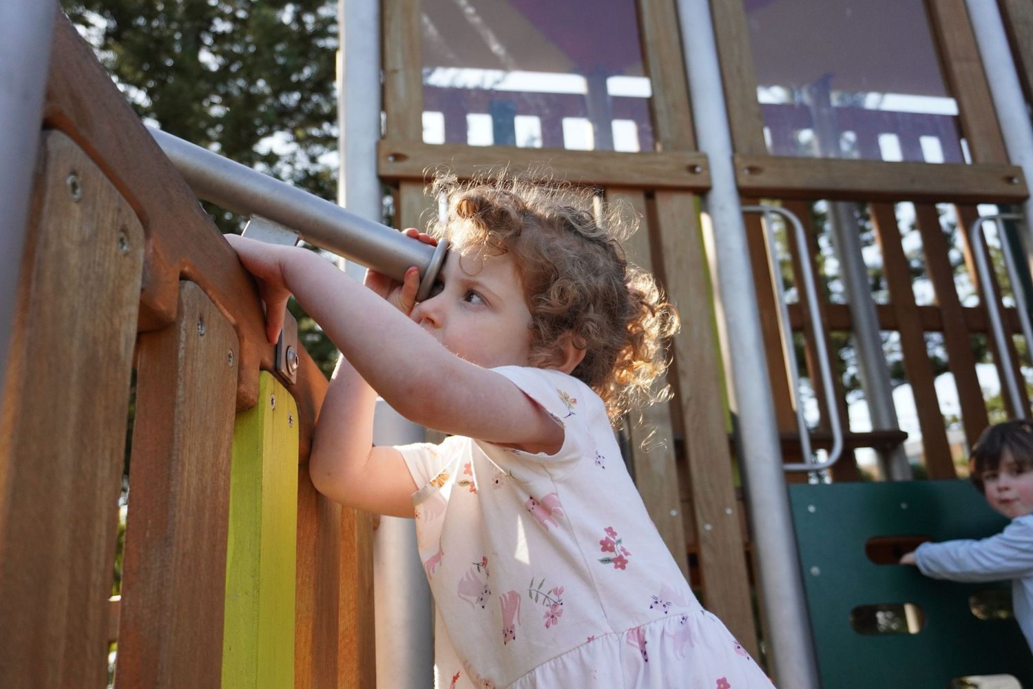 A little girl with curly red hair is playing at a playground. She is looking through a pretend telescope.