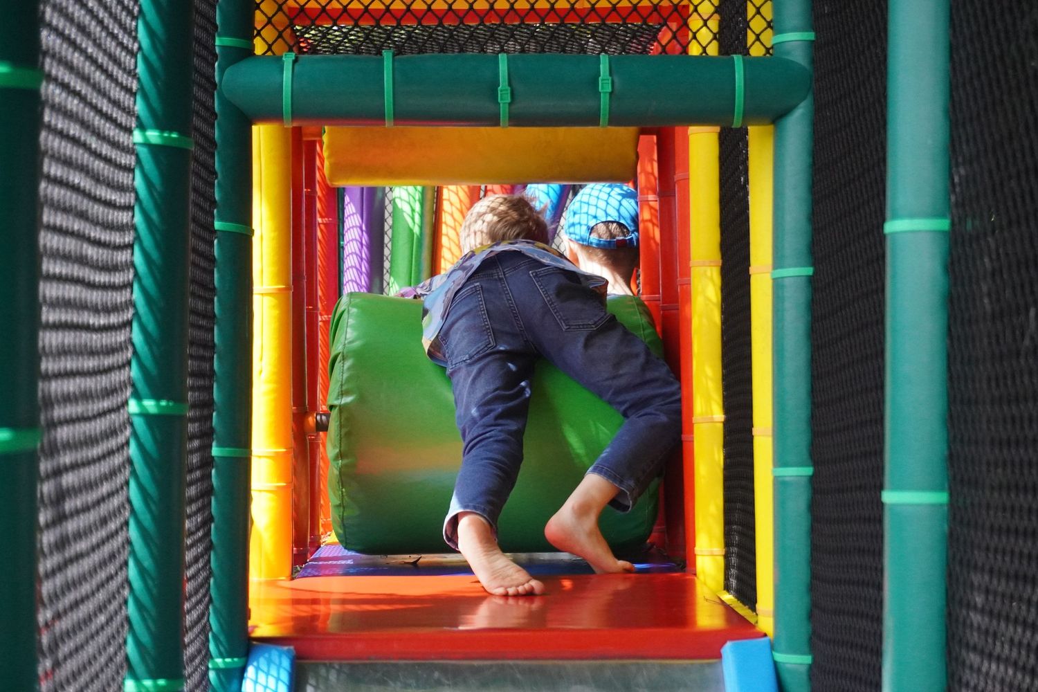 A young boy at an indoor play centre. The photo shows the boy moving away from the camera climbing over a large round green padded wheel.