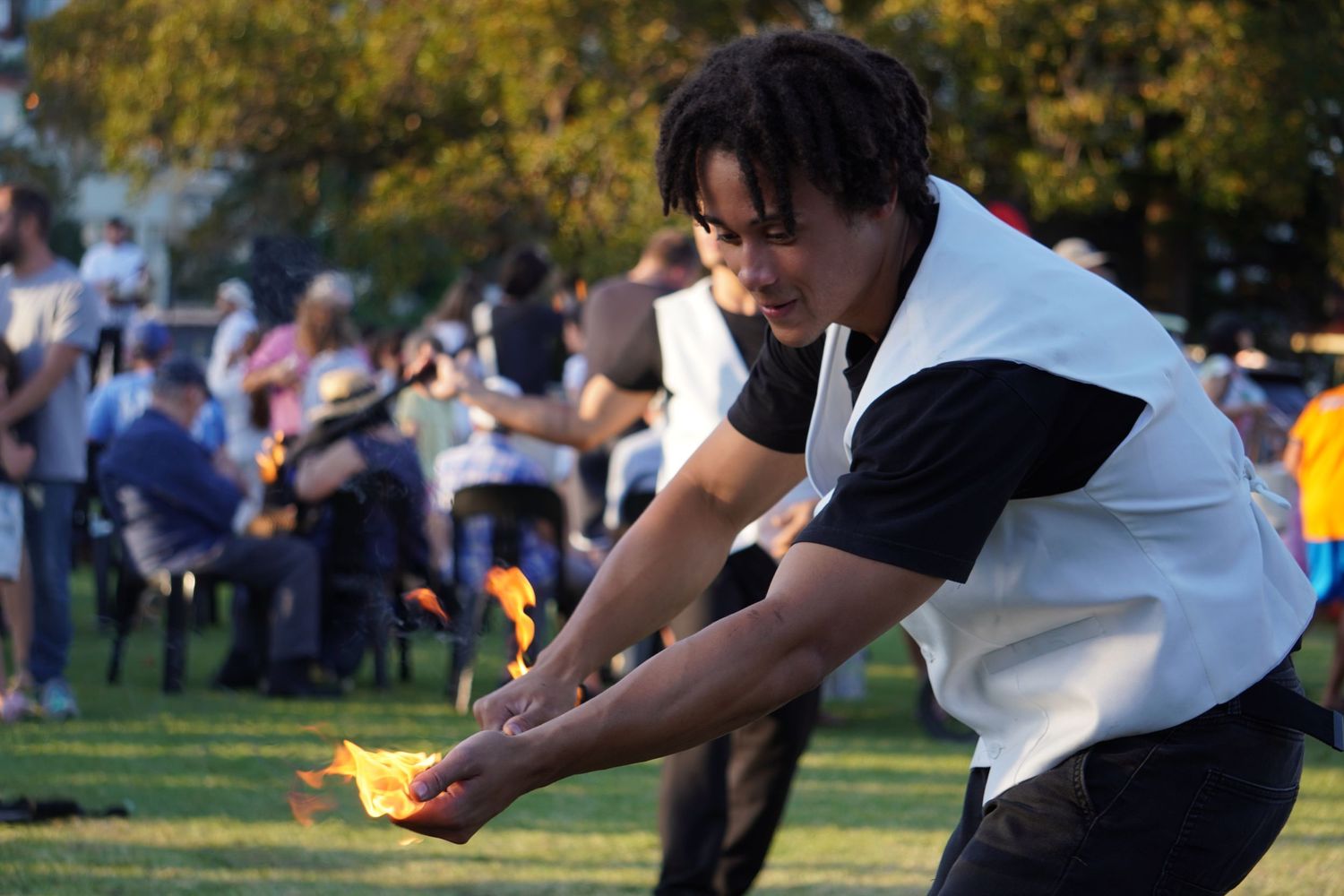 An entertainer at a fair doing tricks with cold fire. He is wearing black clothes with a white vest and is holding the fire in his hands.