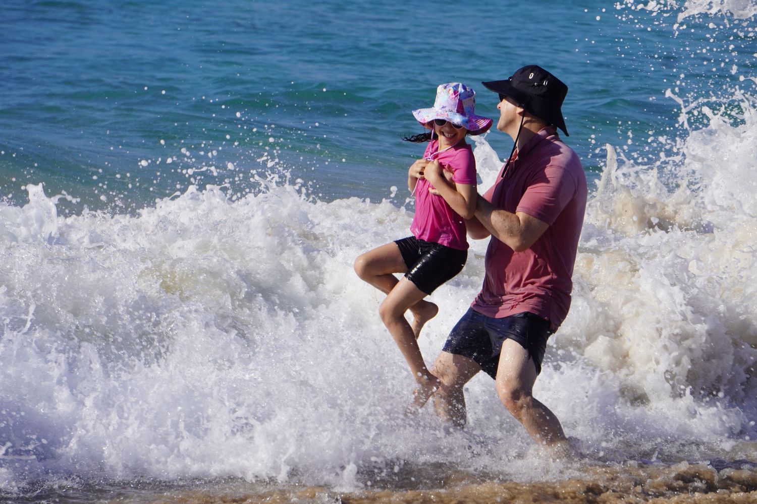 A little girl and her father are at the beach playing in the water. He lifts her as a big wave splashes on them.