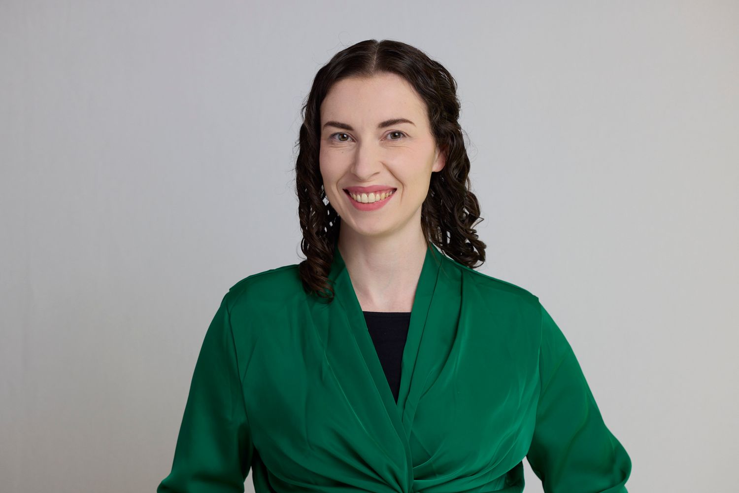 A woman with long brown curly hair smiles at the camera. She is wearing an emerald green satin shirt over a black dress.