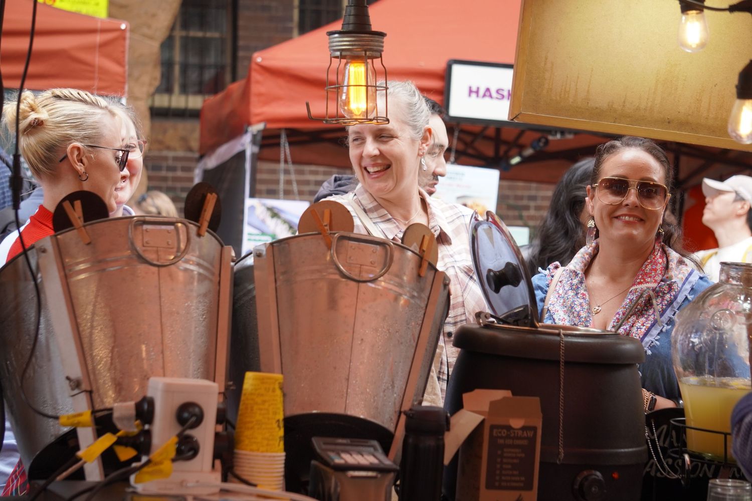 Three friends walk through a busy market street. They are standing at a shop selling mulled wine.