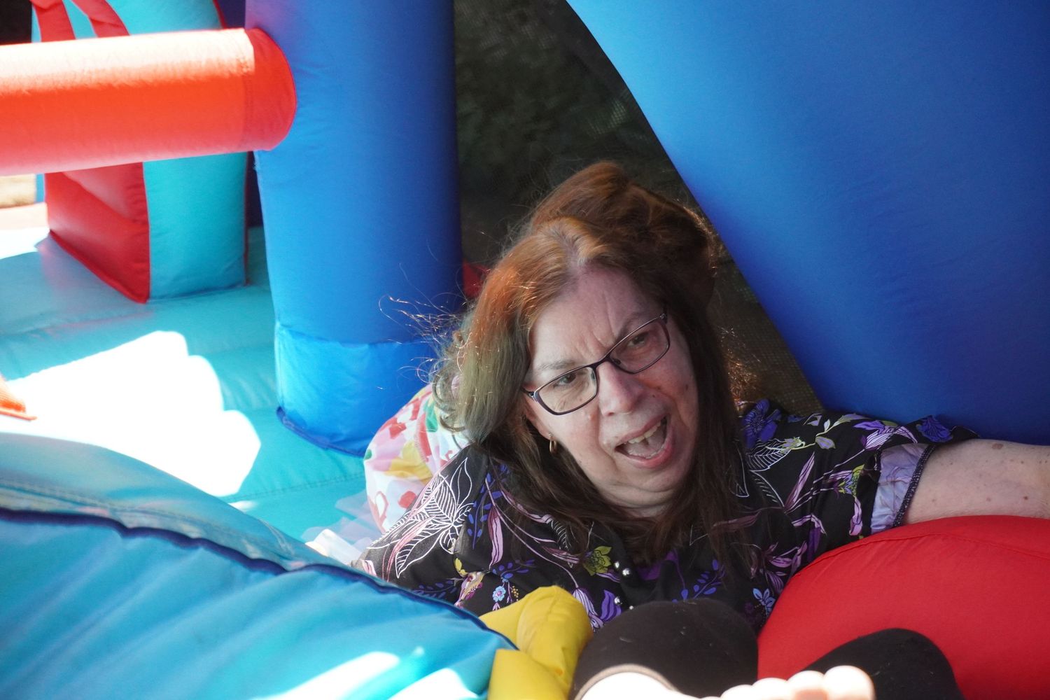 A grandmother is on a blue, red, and yellow jumping castle with her granddaughter. She has fallen and is laughing as she gets up.