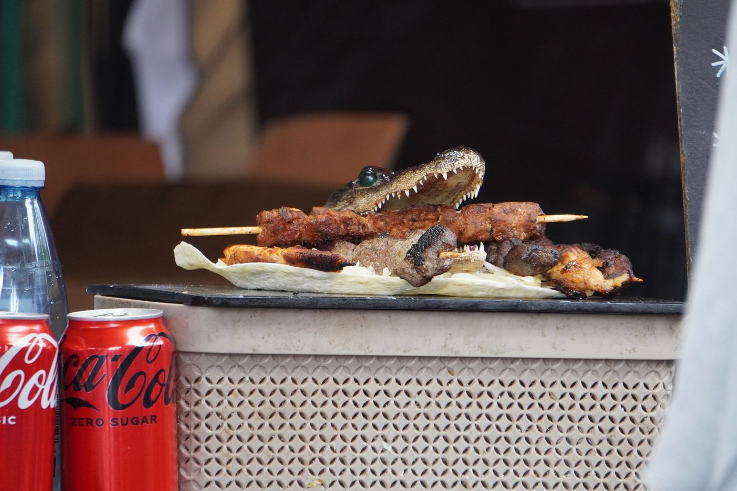 A close-up of a plate of food at a busy food market. It is a small alligator's head. In its mouth is a meat kebab.