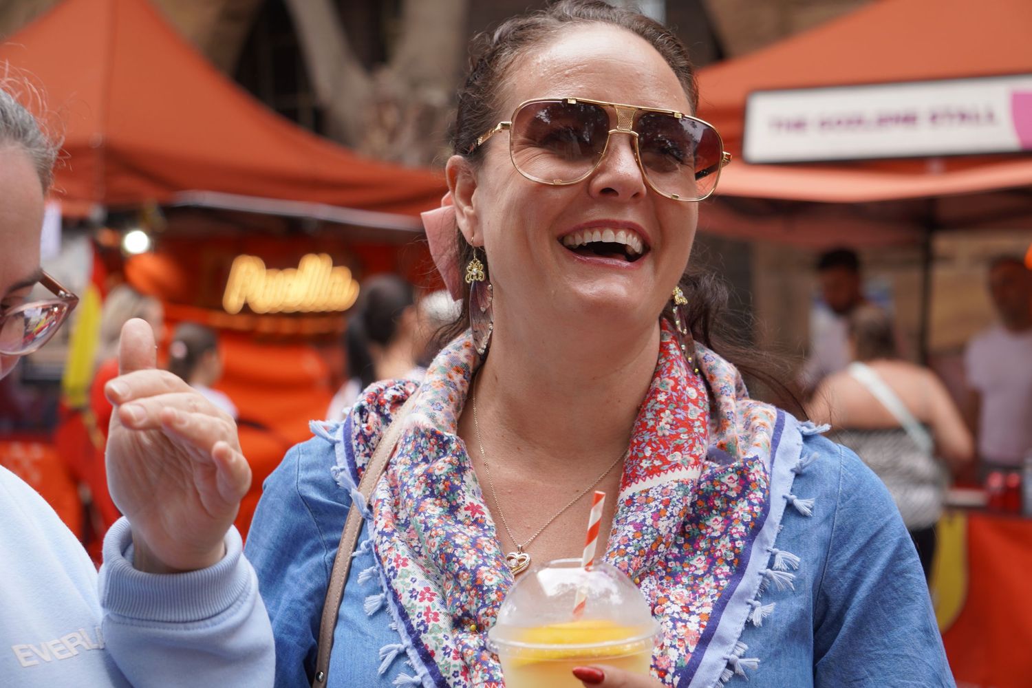 A woman wearing large sunglasses and a denim dress is at a busy market with friends. She is holding a juice and laughing.