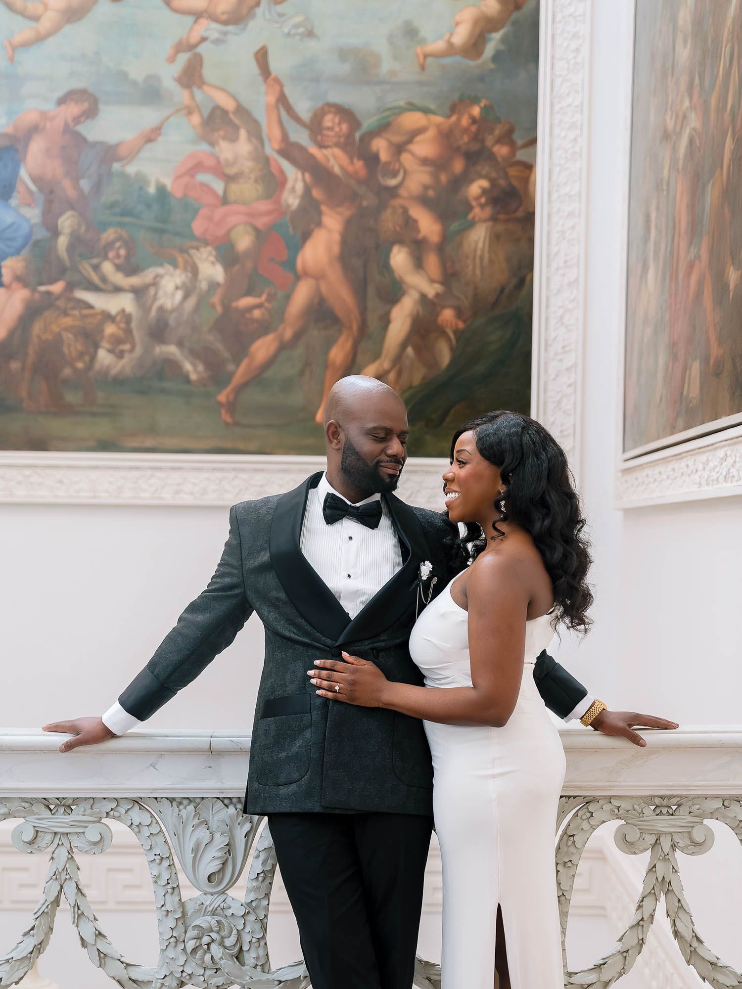 Elegant bride and groom holding hands indoors while smiling against a classical painting background