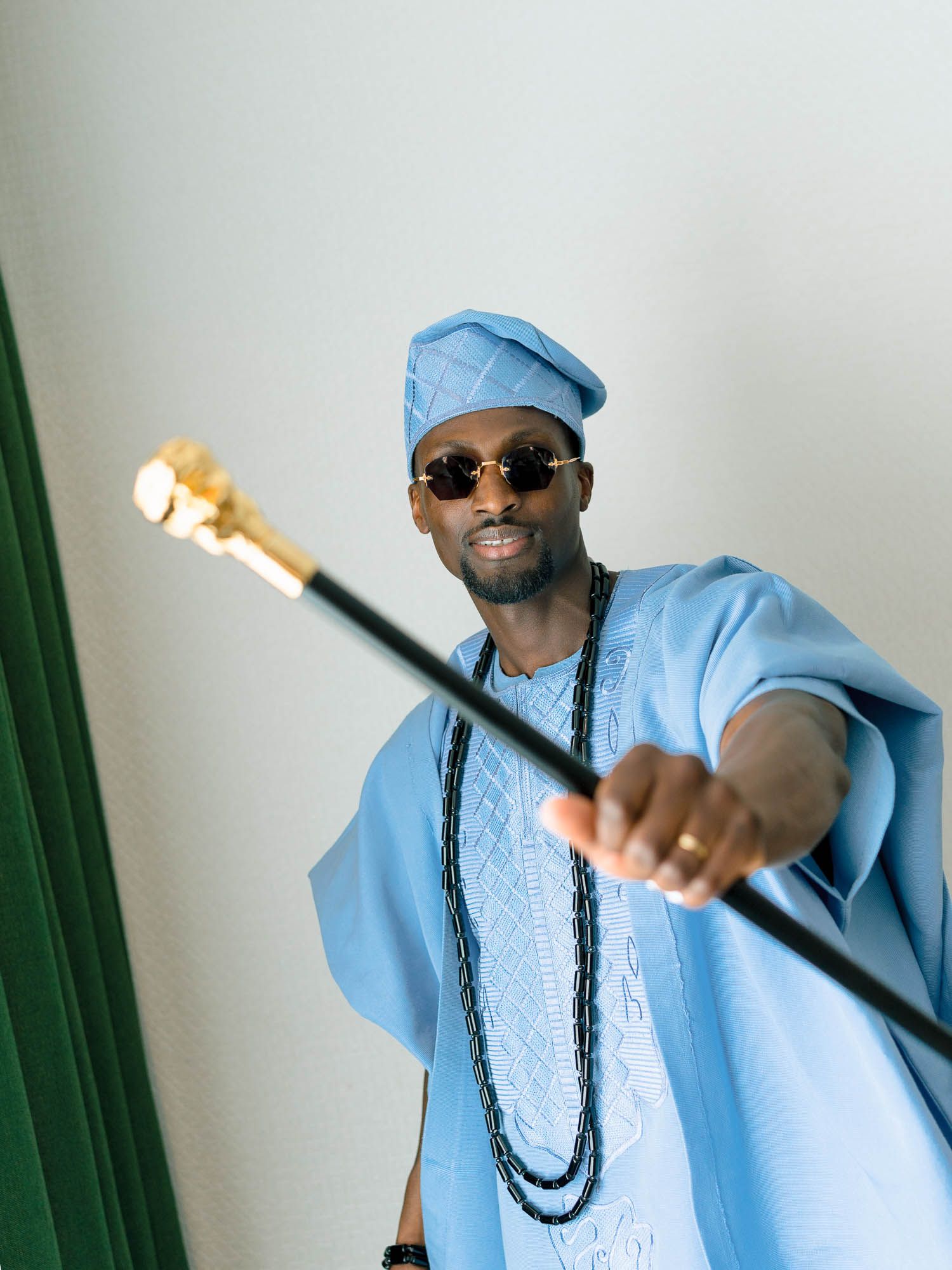 Man posing in a traditional light blue Agbada garment and cap, holding a ceremonial staff.