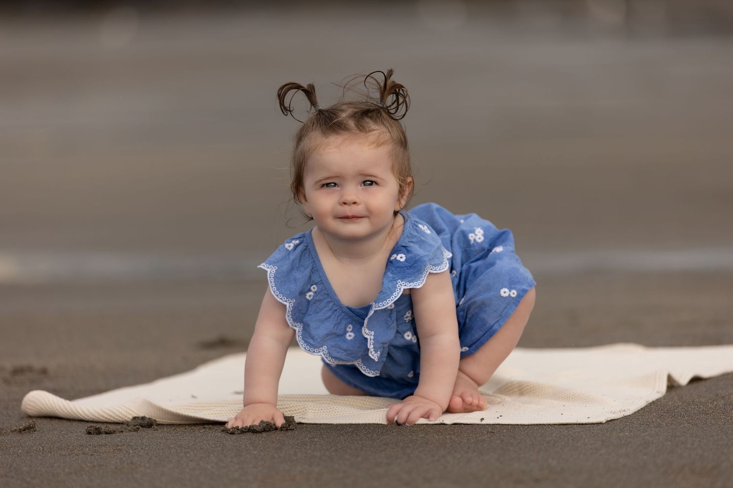 A cute baby girl in a blue dress crawls on a white blanket on a sandy beach.