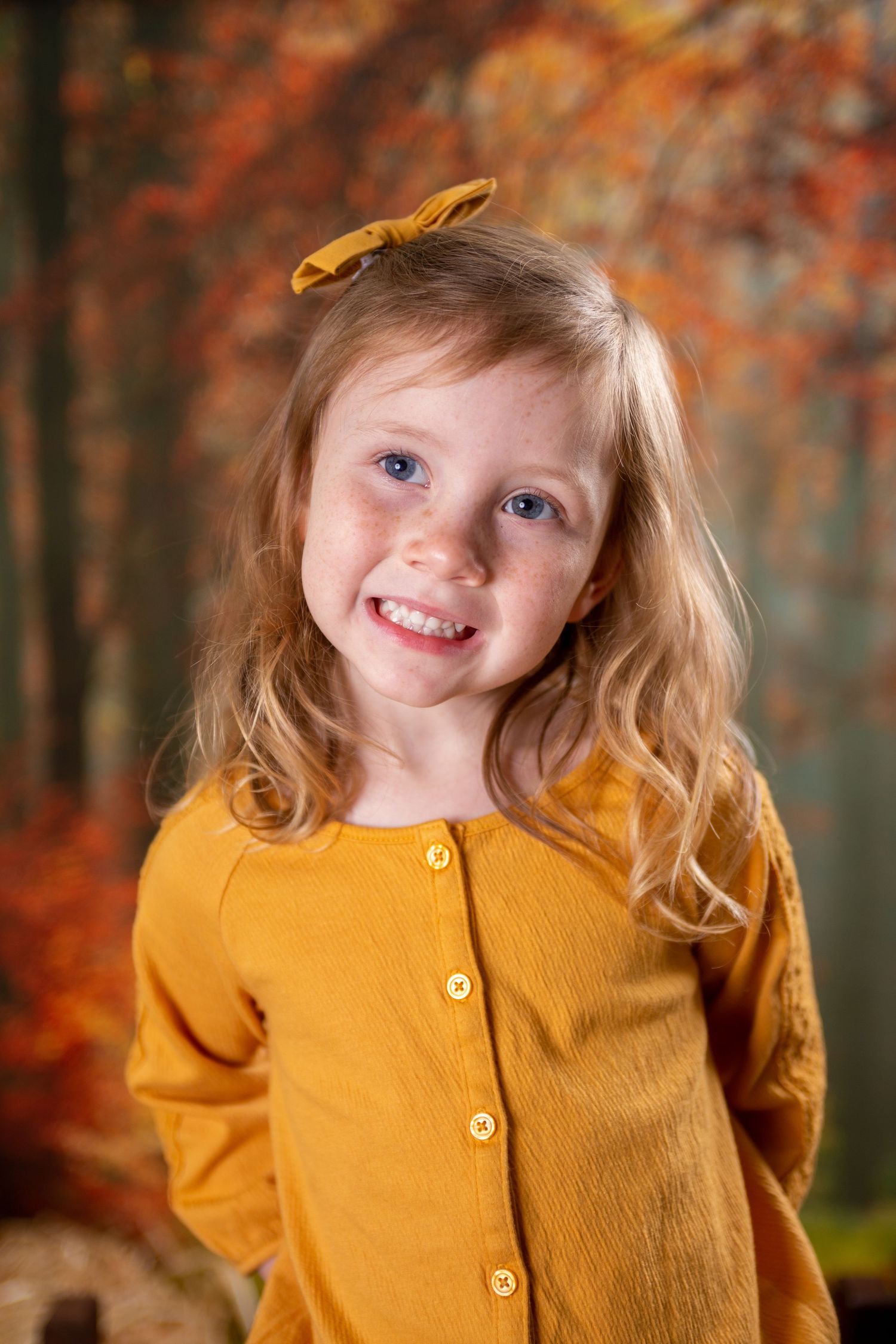 Child wearing a mustard yellow cardigan sweater poses against autumn foliage background with a bright smile.