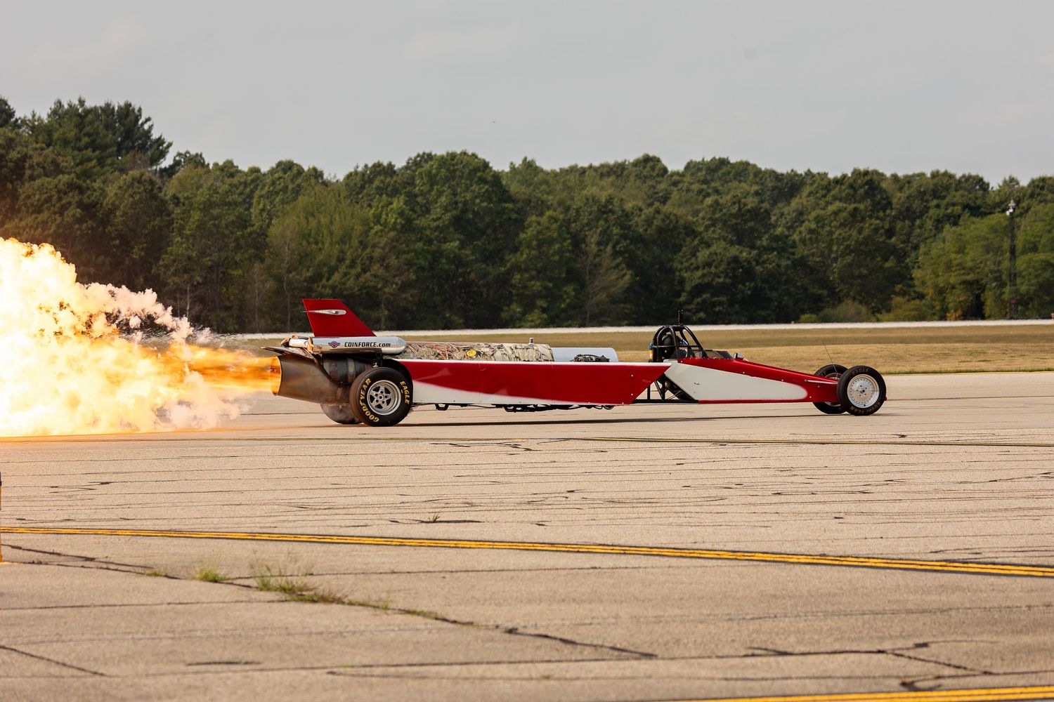 A red jet car performs a high-speed run on a runway with flames shooting from its exhaust during a demonstration.