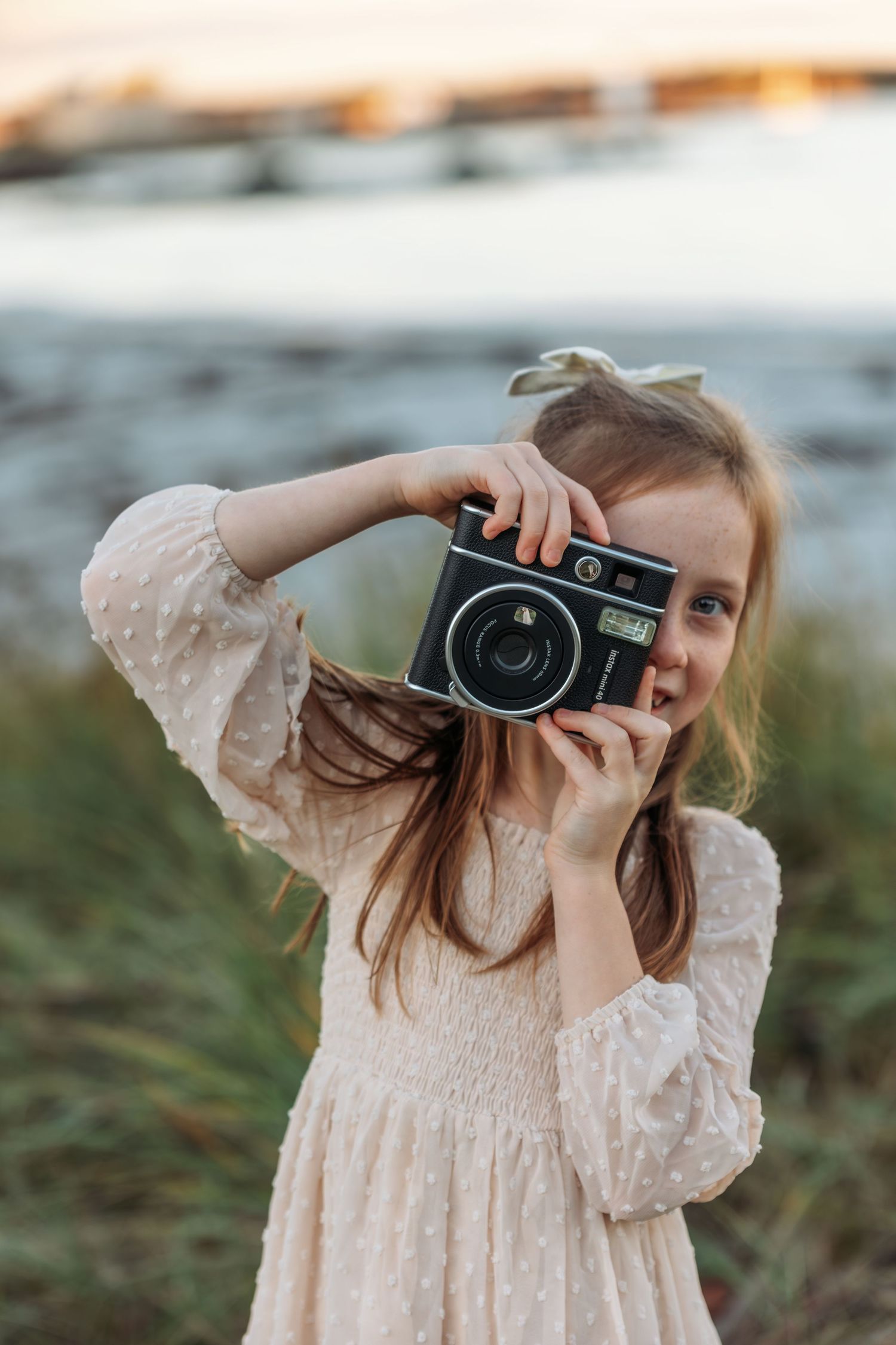 Young photographer in a cream lace dress takes a picture with a vintage film camera at sunset outdoors.