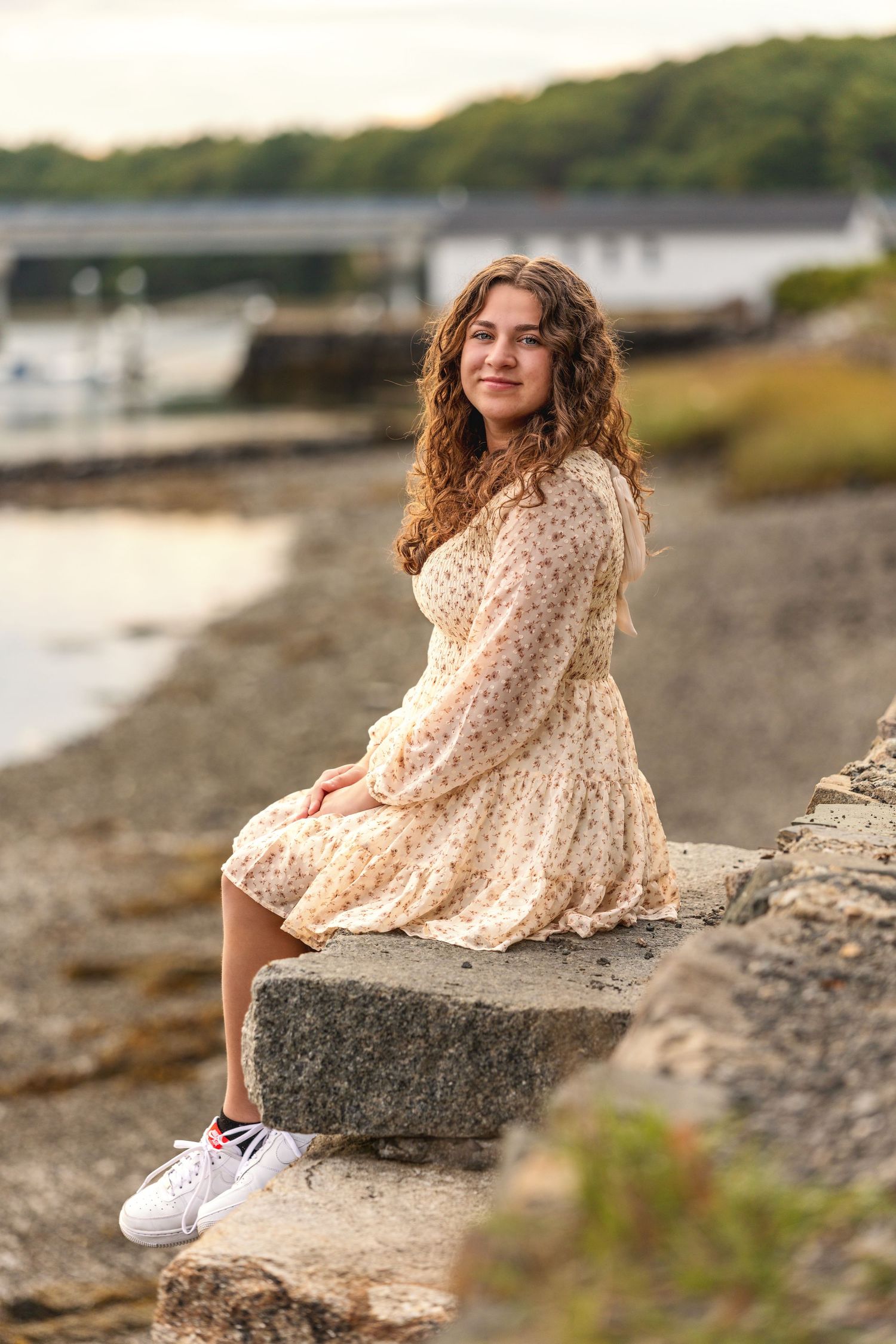 A graduating senior in a polka dot dress sits on coastal rocks at sunset wearing white sneakers.