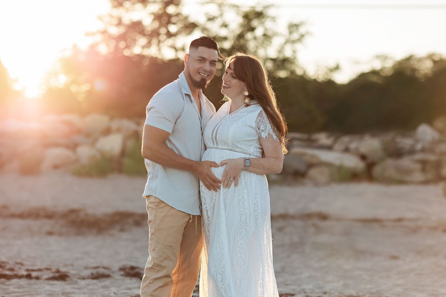A couple embraces on a beach during golden hour sunset as sunlight creates a warm romantic glow around them.