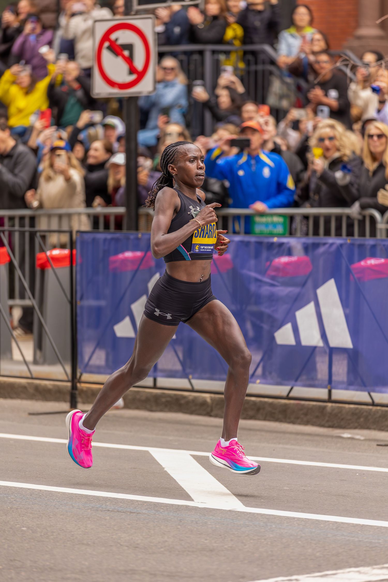 Female marathon runner in pink shoes races past crowds at Boston Marathon, wearing black athletic wear and race bib.