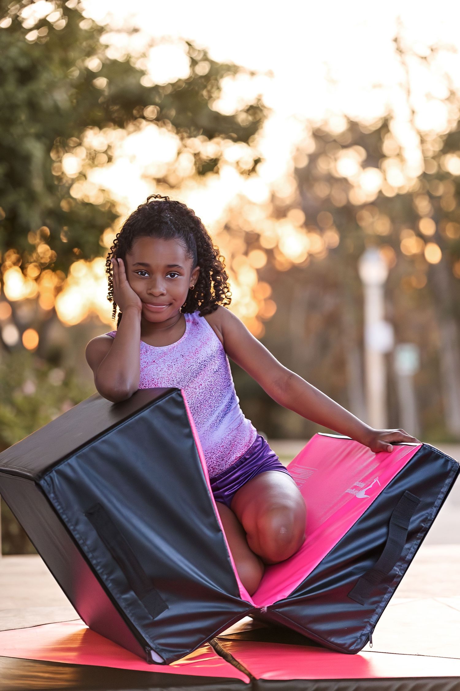 Young girl in purple leotard sitting on a folded gymnastics mat outdoors at golden hour.