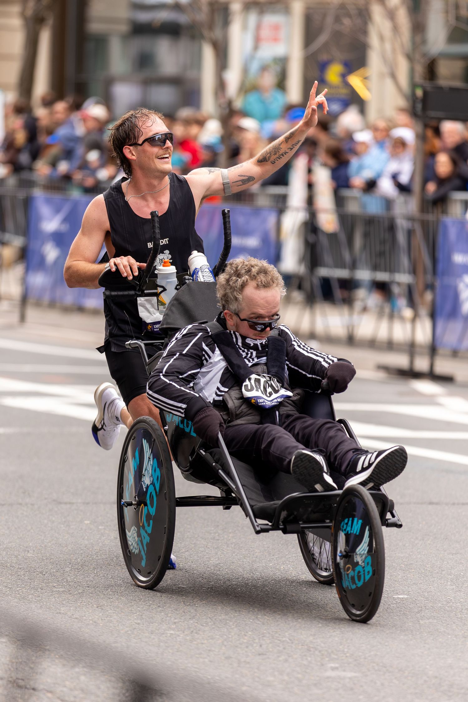 Athlete pushes passenger in racing wheelchair during marathon, waving to crowd lining urban street course.