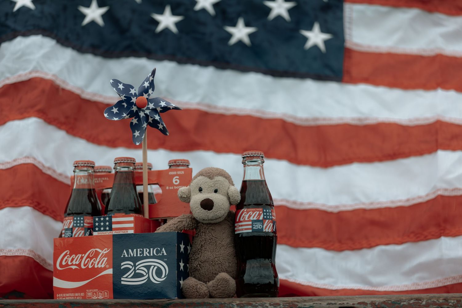 Coca-Cola America 250 anniversary bottles with a teddy bear and pinwheel in front of an American flag.