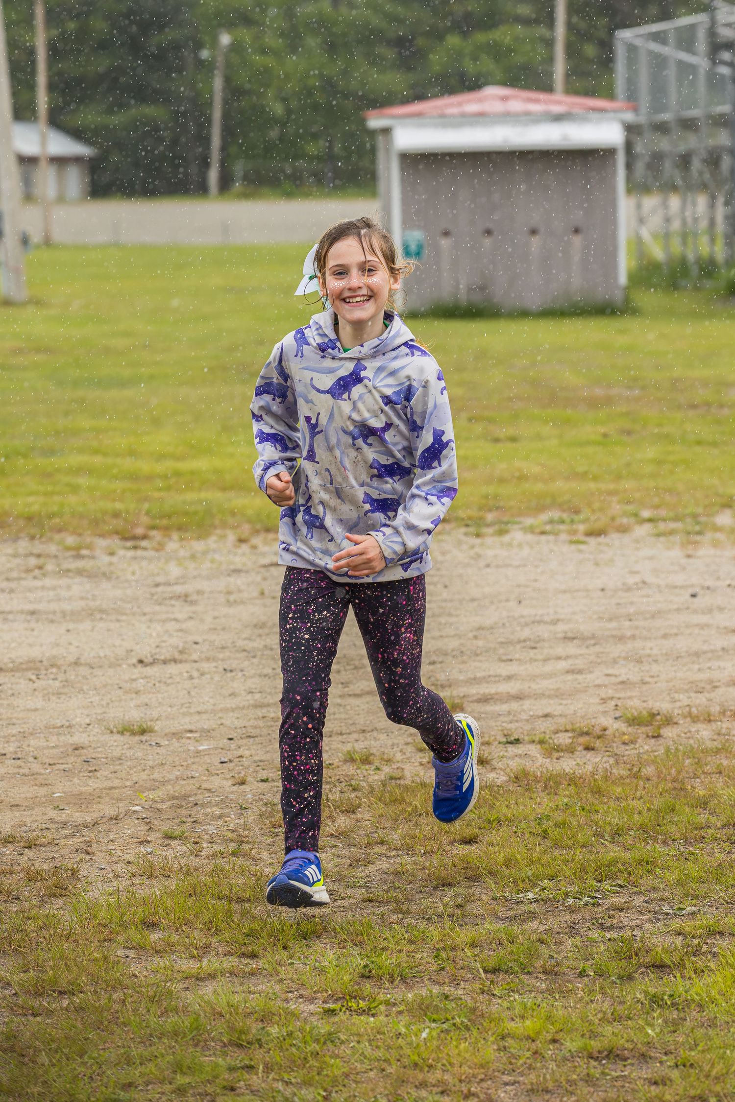 A smiling young girl in a blue tie-dye hoodie and leggings runs happily across a grassy field in the rain.