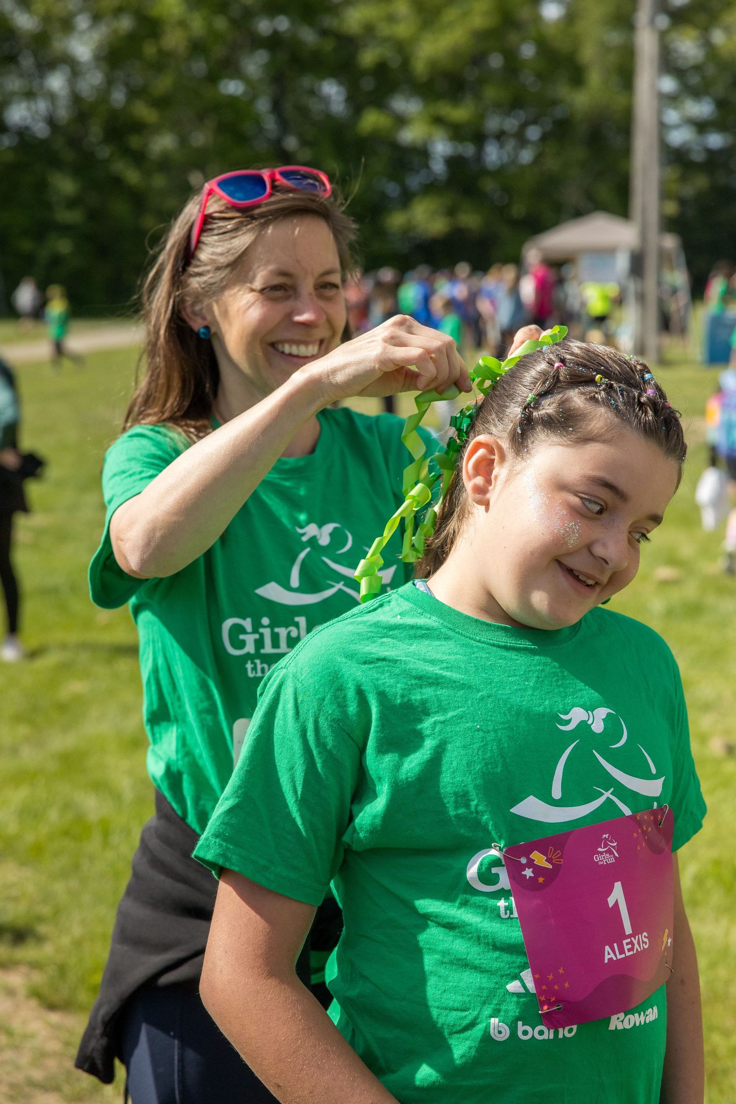 Woman braiding a girl's hair at a Girls on the Run outdoor event, both wearing green t-shirts.