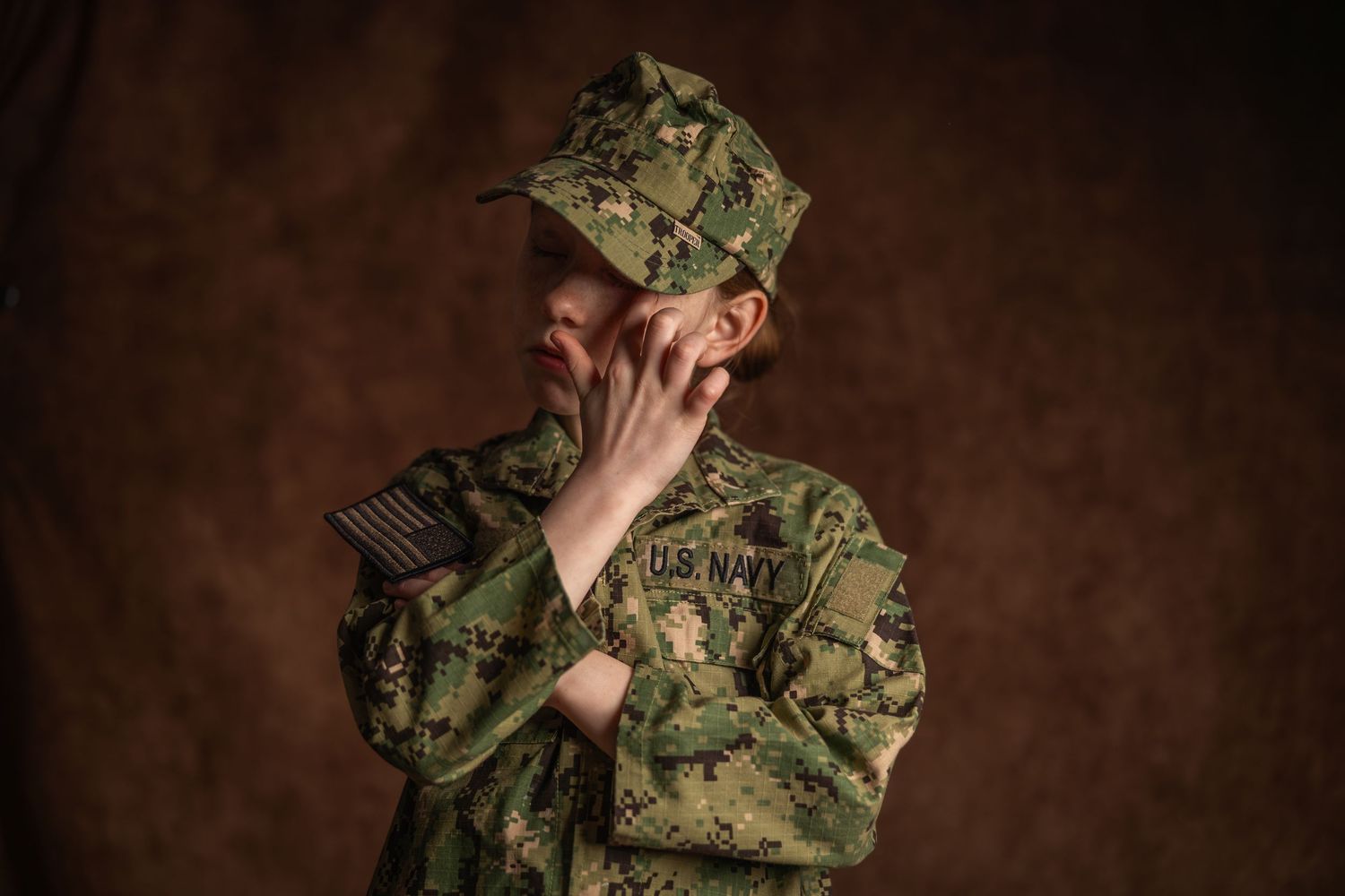 Female U.S. Navy soldier in camouflage uniform and cap posing thoughtfully against brown background.