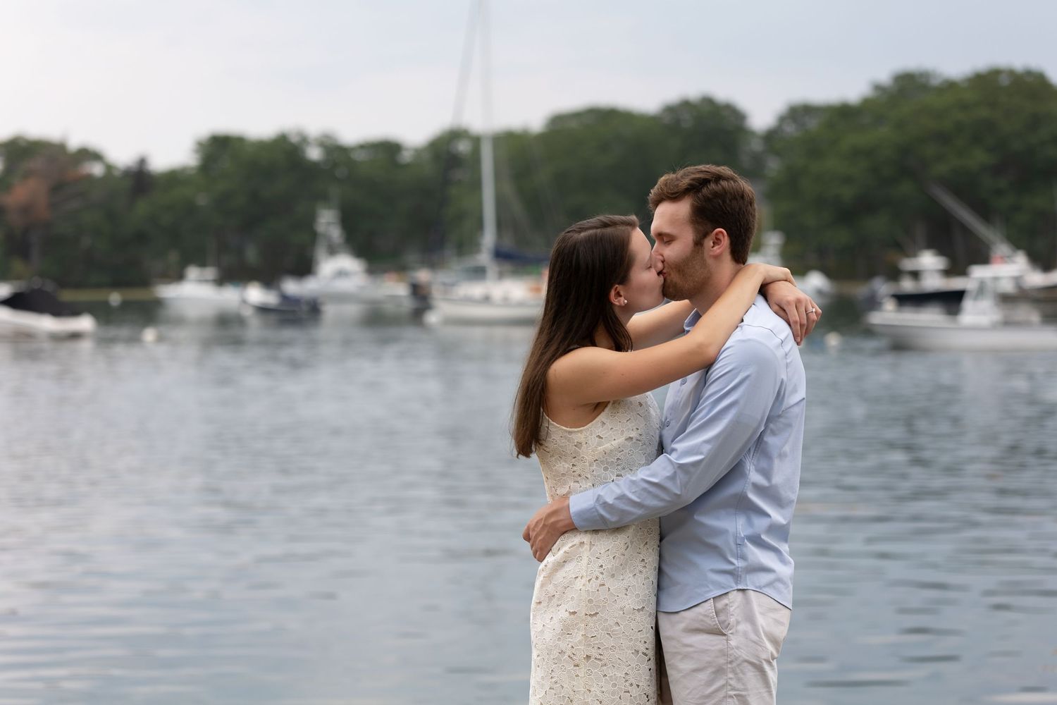 A romantic couple embraces and kisses by a scenic waterfront Fisherman's Path, York, ME with boats anchored in the harbor on a cloudy day.