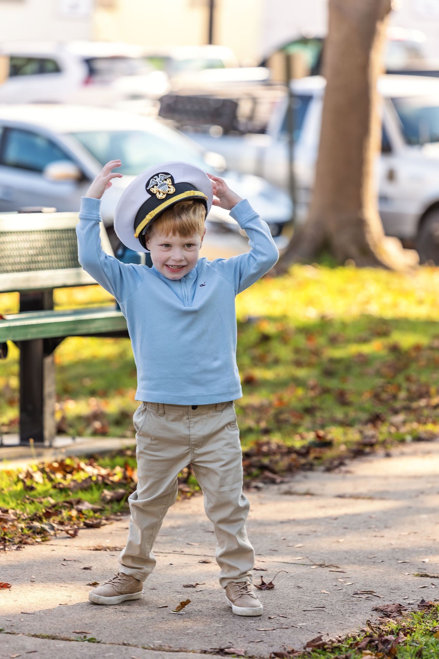 Smiling young boy wearing a white captain hat in a park, dressed in a blue sweater and khaki pants.