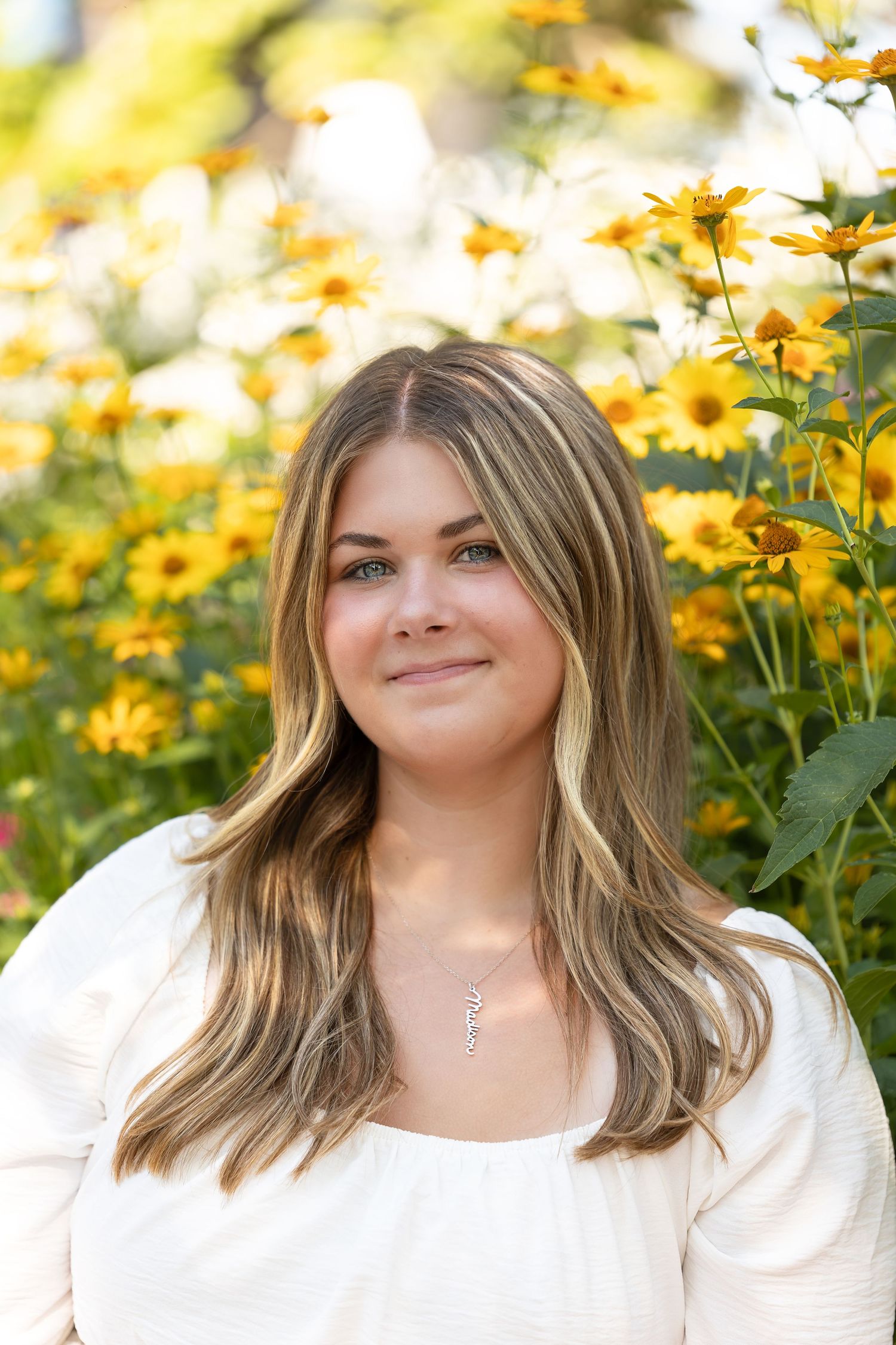 Young woman with highlighted hair smiles in front of vibrant yellow flowers, wearing a white top and delicate necklace.