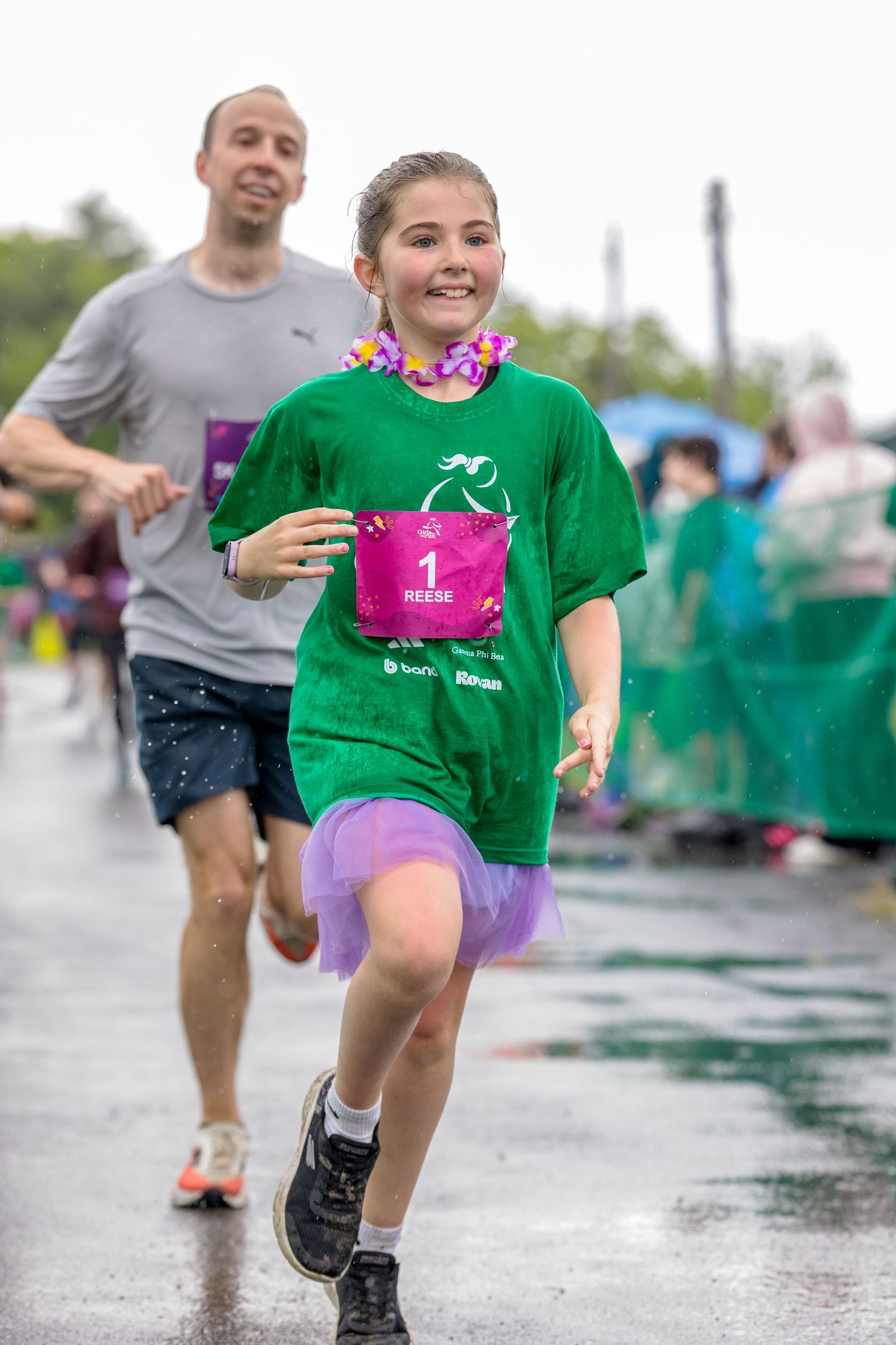 Young girl in green shirt and purple tutu runs race wearing bib number 1, smiling as man follows behind on wet course.
