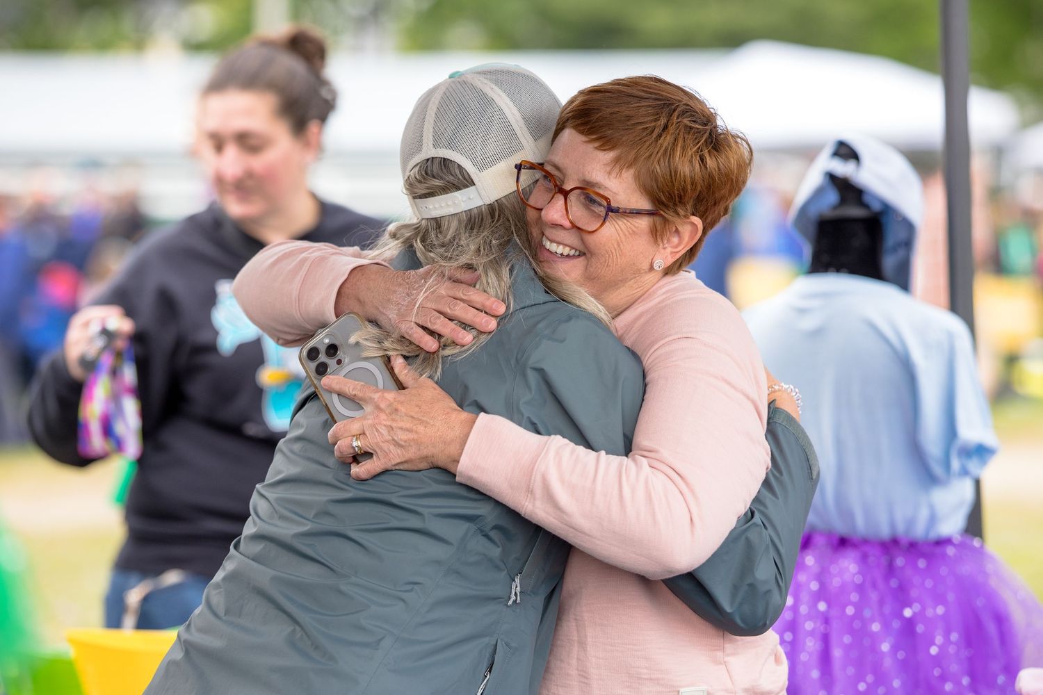Two women share a warm hug at an outdoor festival, smiling joyfully amid a colorful crowd.