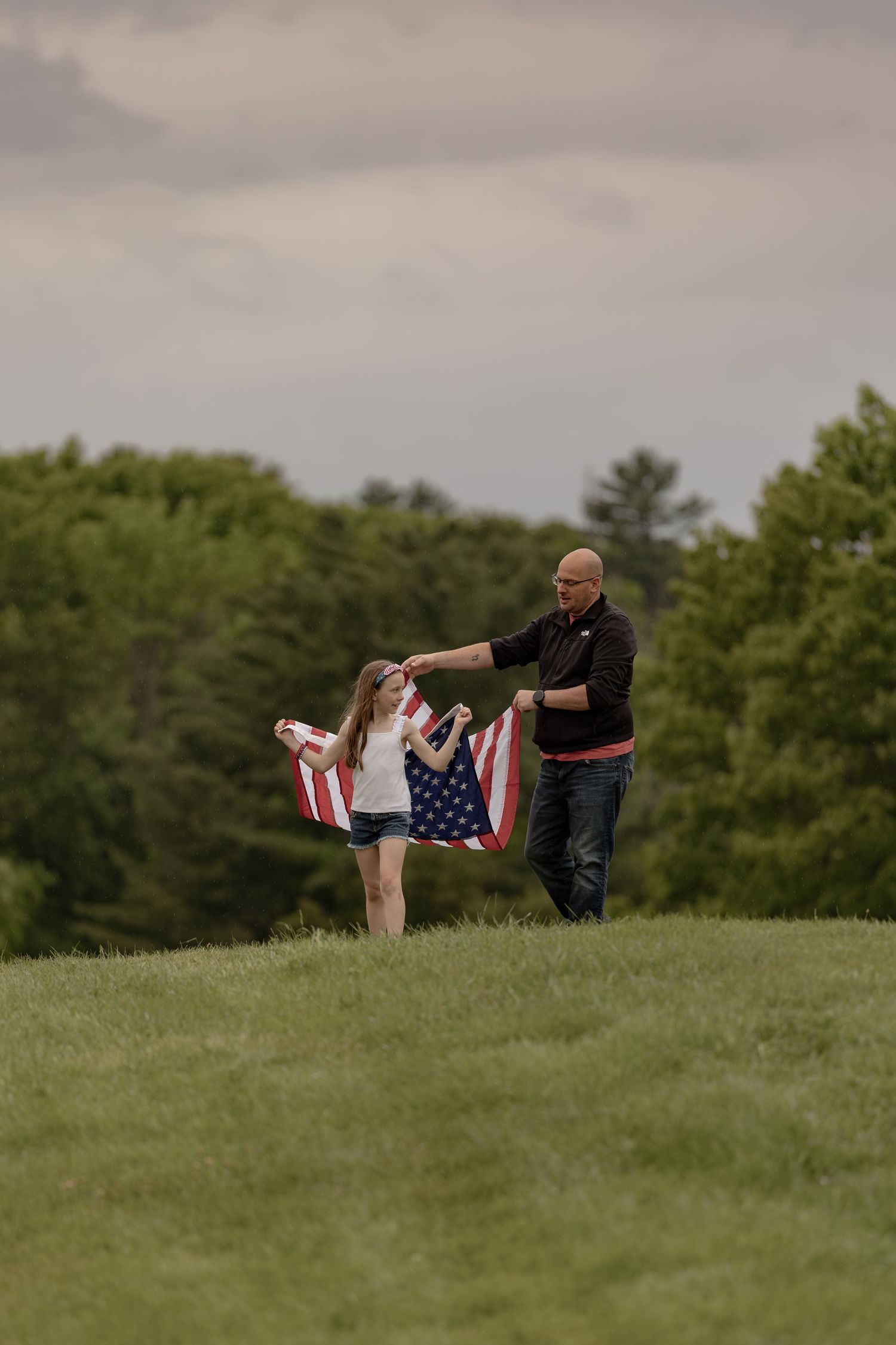 A man and young girl hold an American flag on a green hillside with trees in the background.