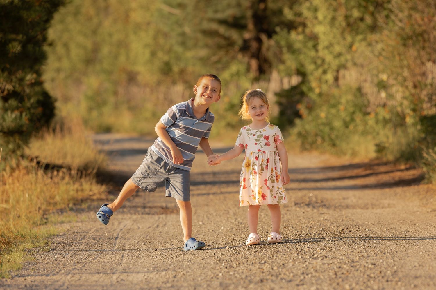 Two children running joyfully on a sunny dirt road surrounded by trees, a boy in stripes leading a toddler girl in a floral dress.