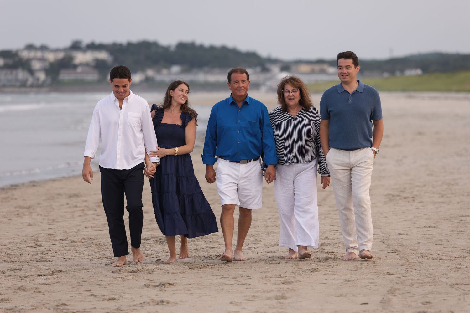 Family of five walking together on a sandy beach, smiling and enjoying a casual seaside stroll on an overcast day.