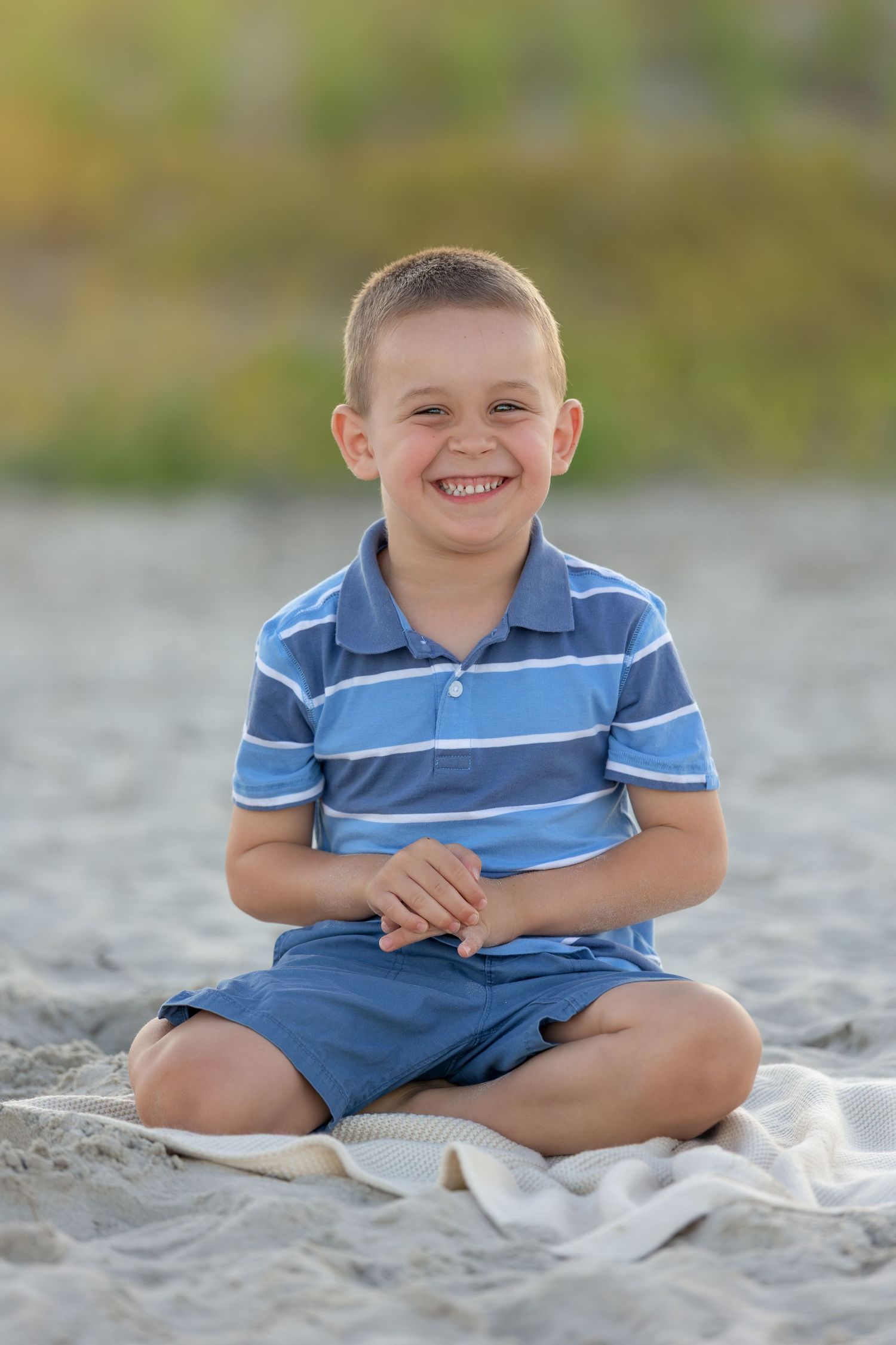 Smiling young boy in blue striped polo shirt sitting cross-legged on sandy beach blanket outdoors.