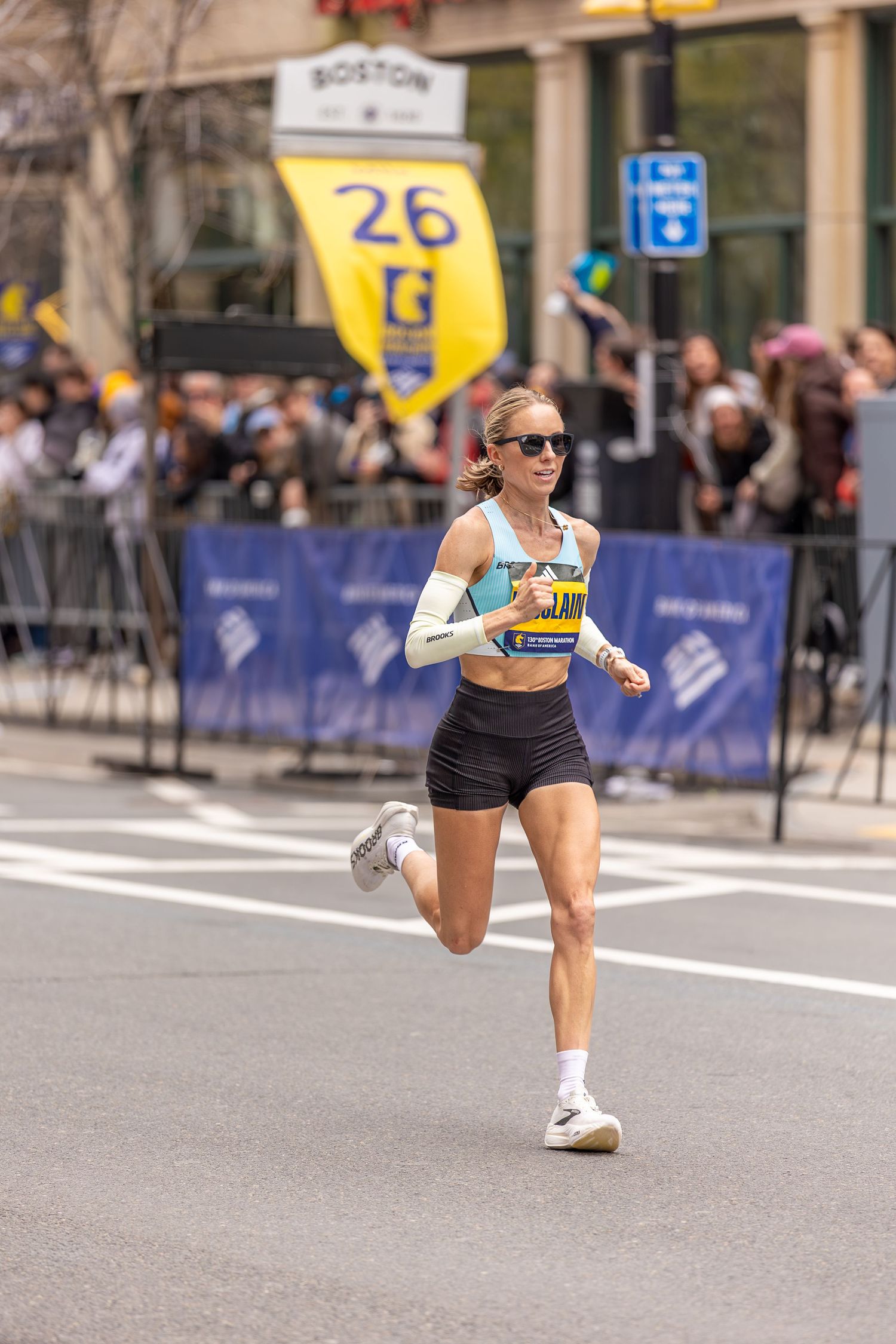 Female runner competing near mile 26 marker at the Boston Marathon, wearing sunglasses and race bib.