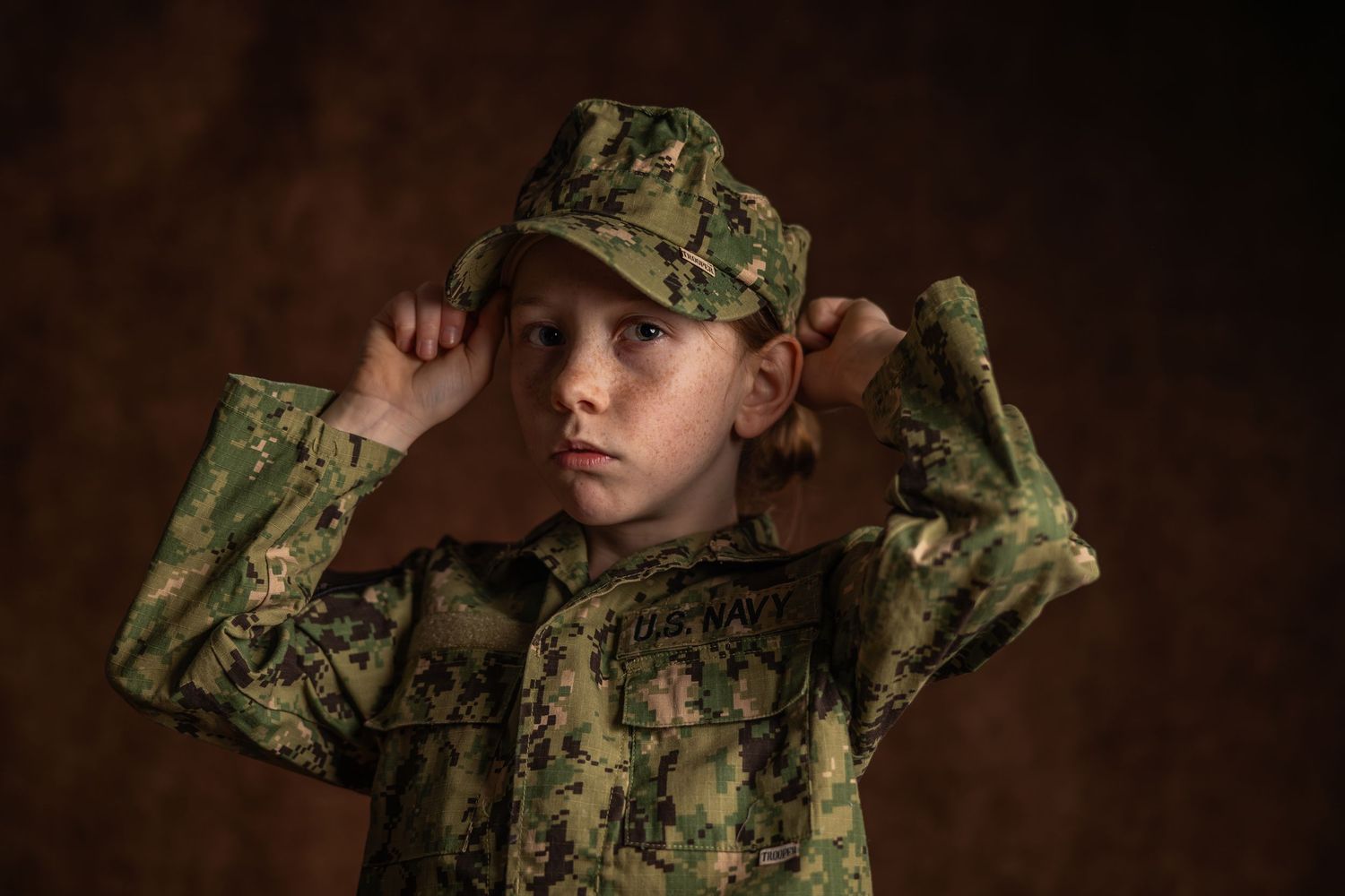 Young child wearing oversized camouflage military uniform and cap, posing seriously against dark brown background.